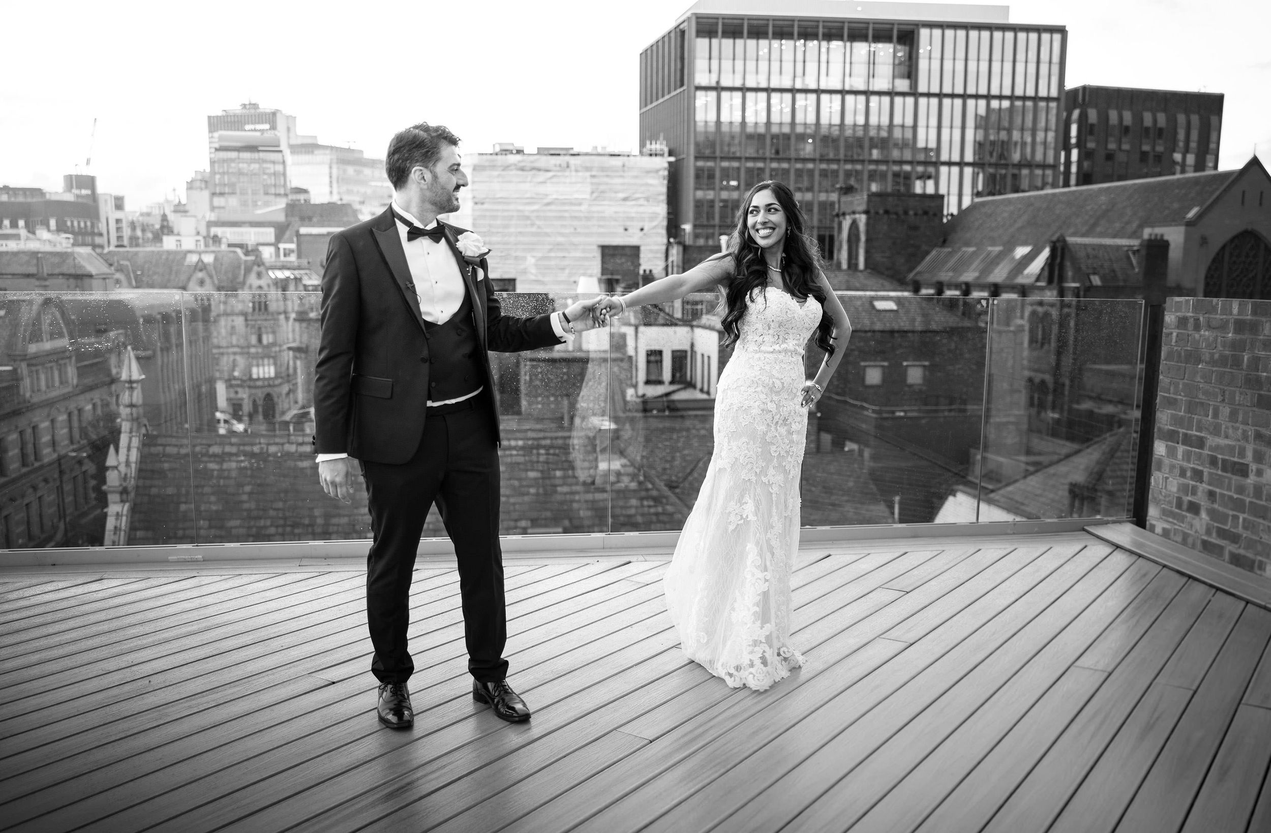 Black and white photo of bride and groom holding hands on rooftop terrace with city skyline backdrop.
