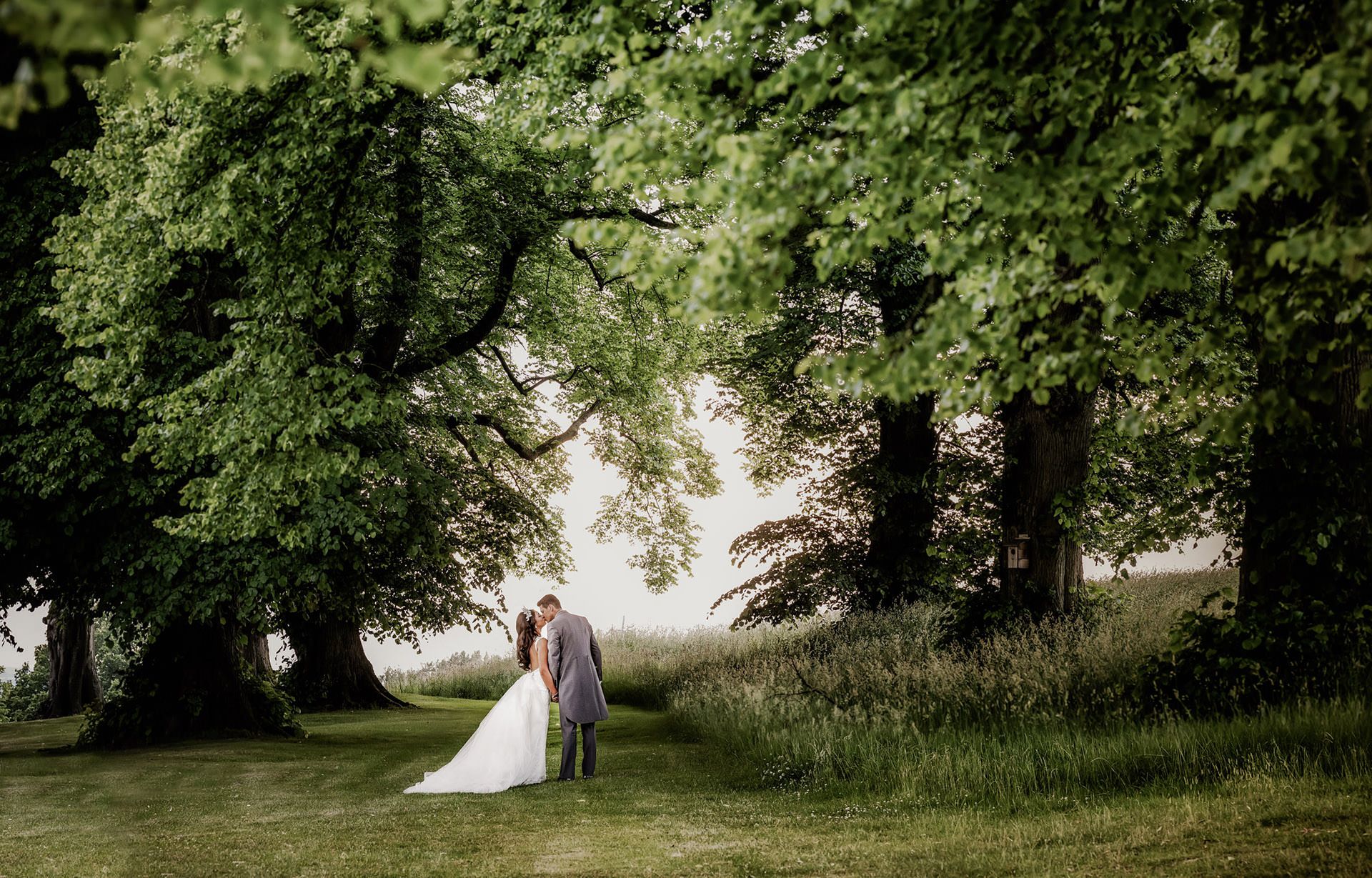 A bride and groom share a romantic moment beneath lush green trees in a sunlit garden setting.
