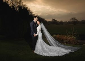 Bride in lace mermaid gown with long veil and groom in navy suit pose romantically at dusk on lush green grounds.