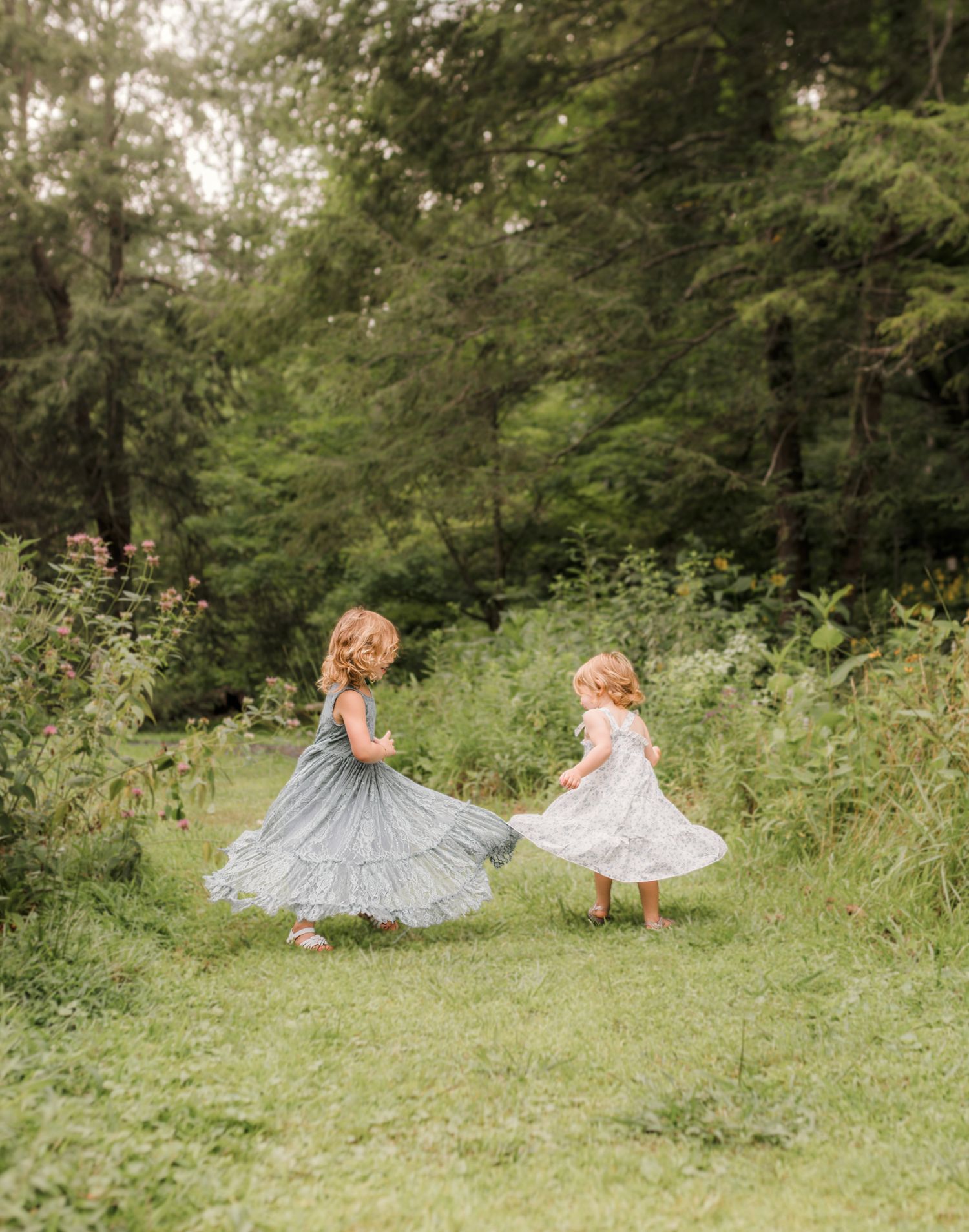 Two young girls in flowy dresses twirl together in a lush green meadow surrounded by tall trees.