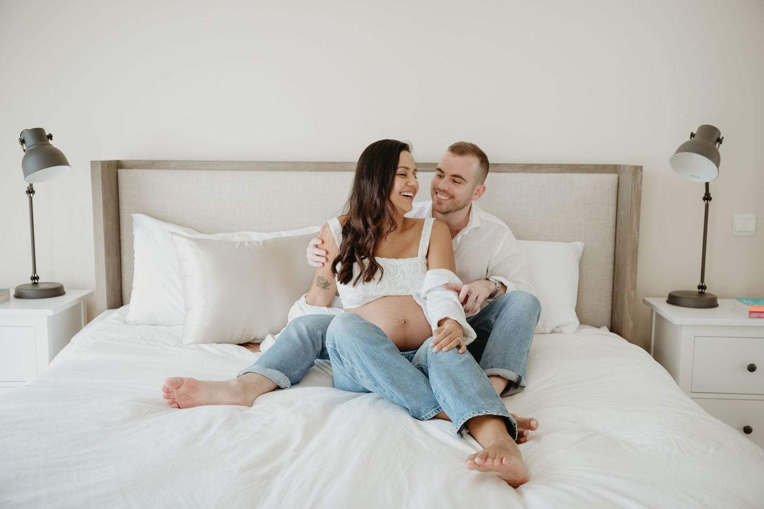 Happy couple sitting on a white bed, smiling and embracing in a bright, cozy bedroom with nightstands.