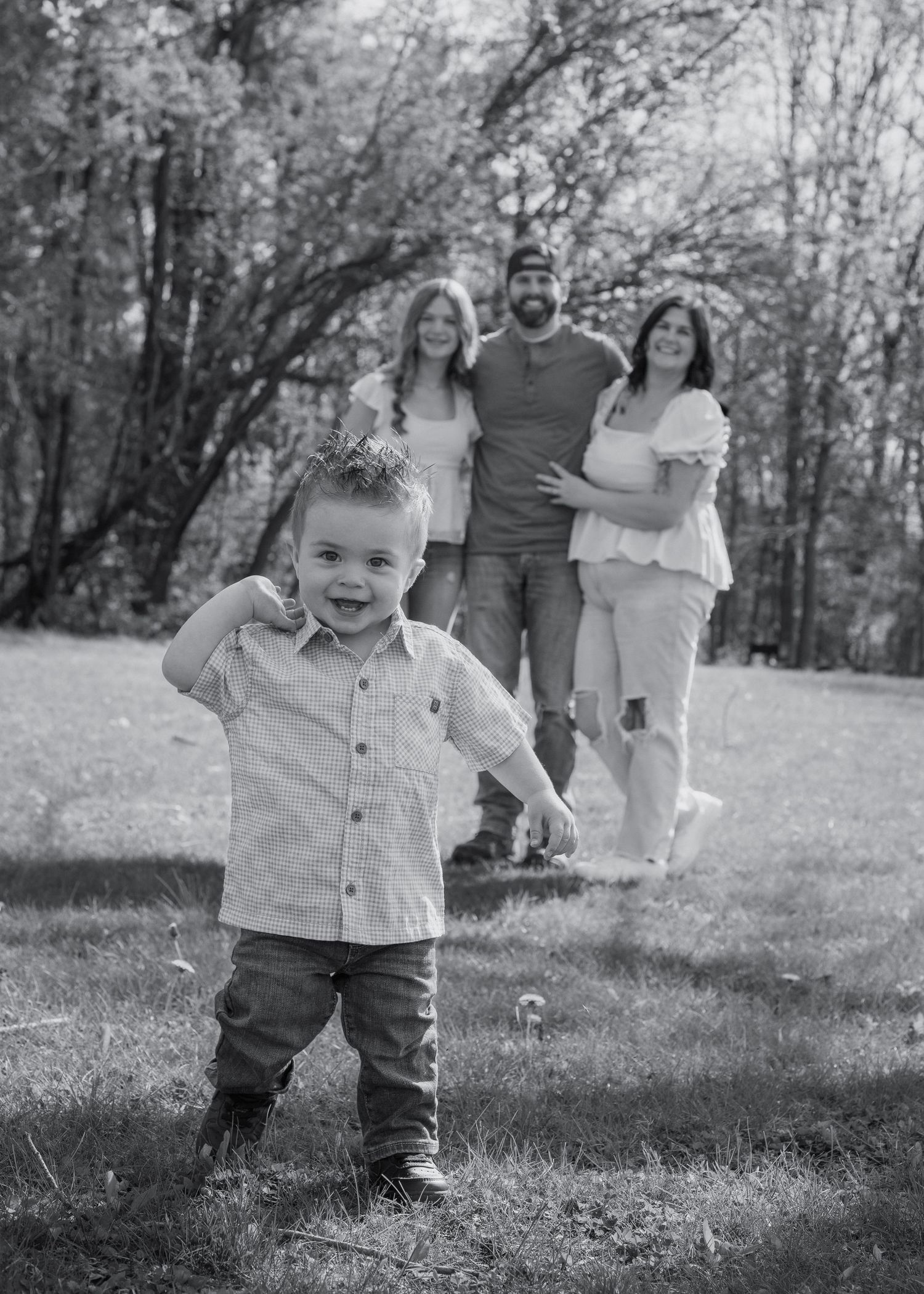 Smiling toddler poses in foreground while family of three stands in blurred background, black and white.