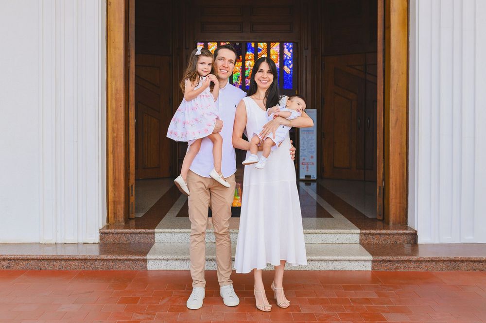 Happy family of four posing outside a church, parents holding two young children, dressed in elegant white and neutral outfits.
