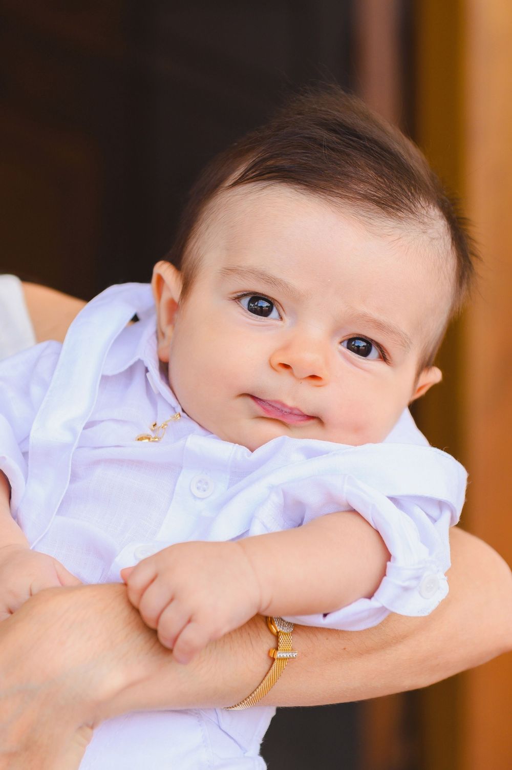 A cute baby with dark hair and blue eyes wearing a white outfit, being held by an adult wearing a gold bracelet.