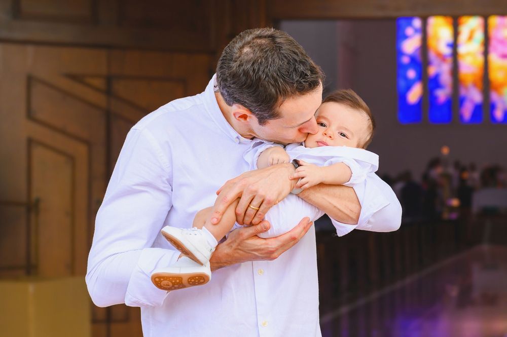 A father tenderly kisses his baby during a church baptism ceremony, both dressed in white.