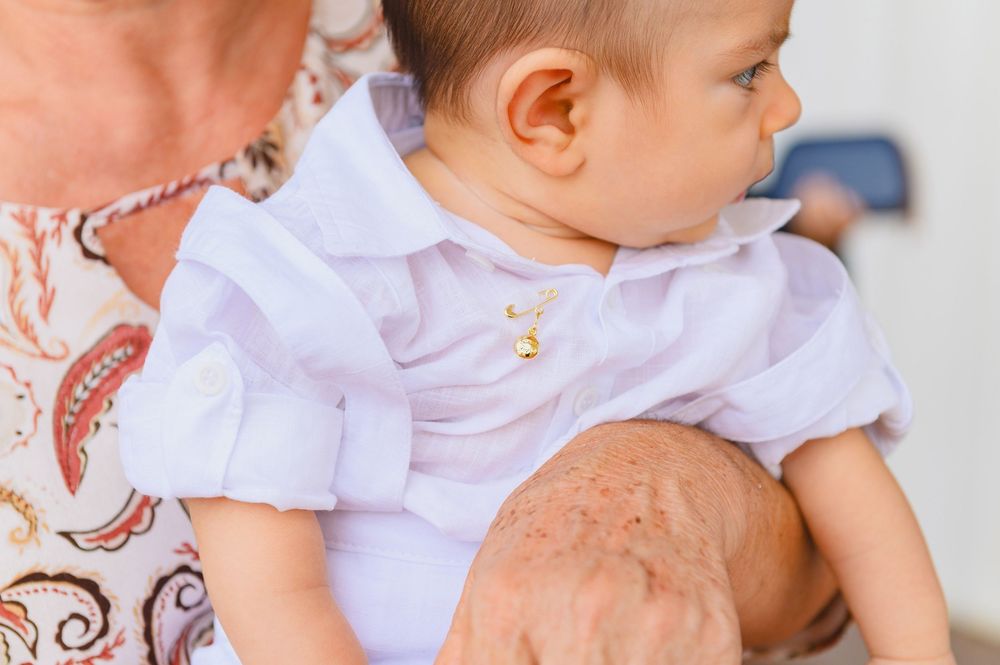 Baby in white christening outfit with gold cross necklace held by adult in floral dress.
