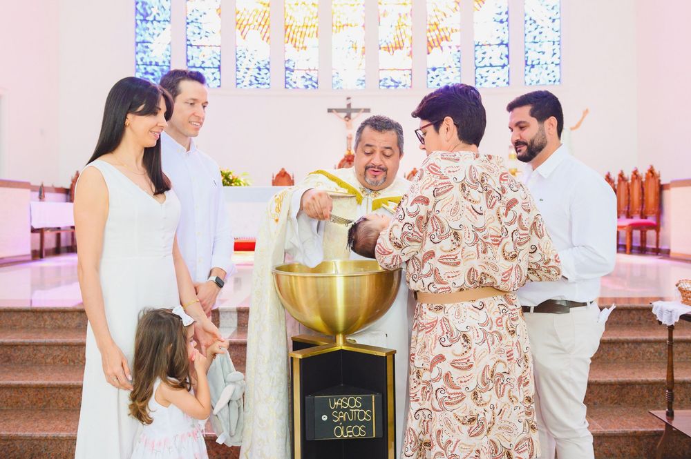 Catholic baptism ceremony with priest, family, and godparents gathered around golden font in white church.