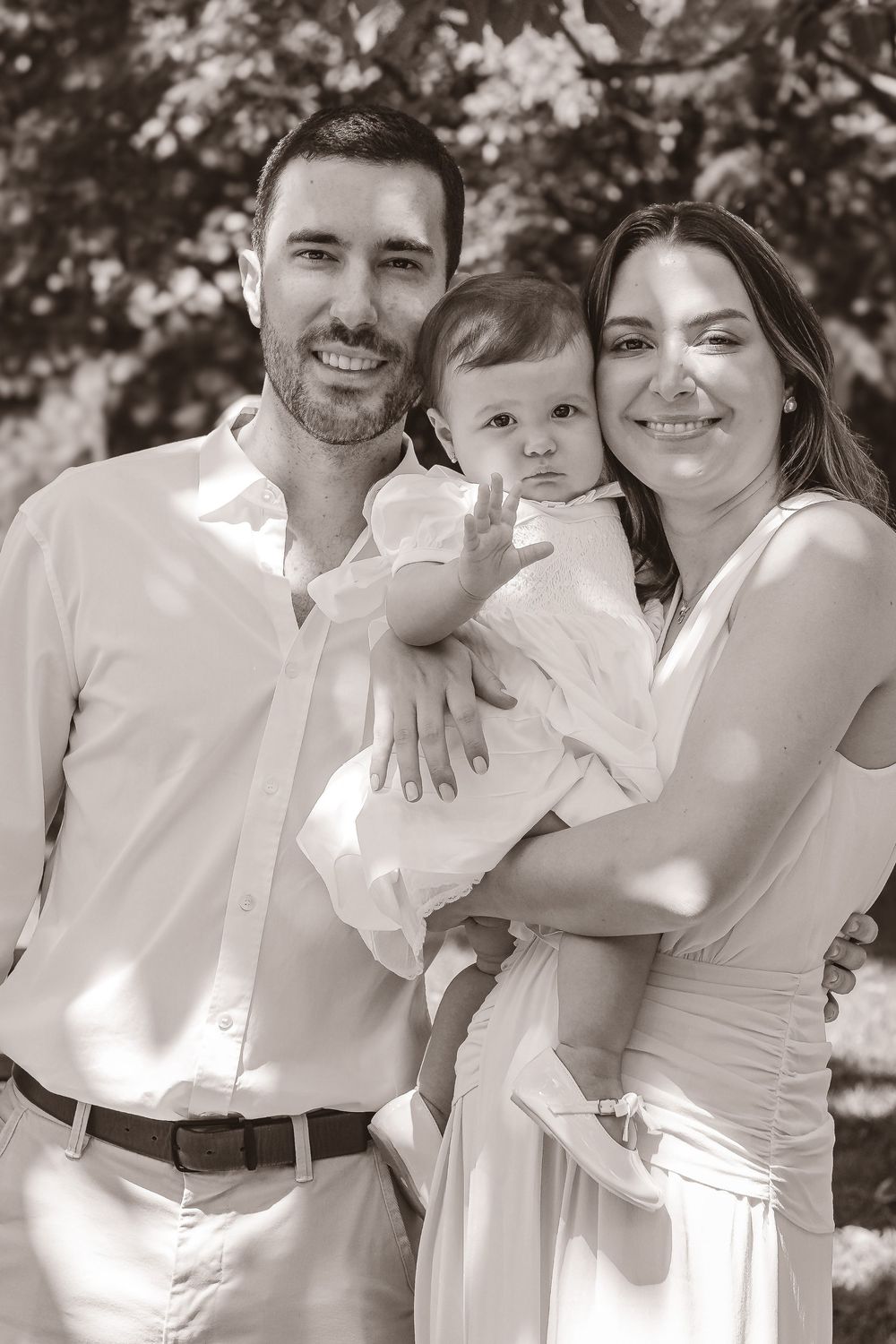 Black and white portrait of smiling couple holding baby in white outfit outdoors with trees in background.