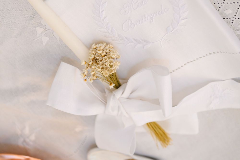 White wedding candle with gold floral ring and satin ribbon bow on embroidered fabric background.