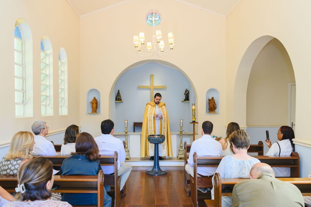 Priest in gold vestments leads a small congregation in a bright, intimate chapel with a cross and candles at the altar.