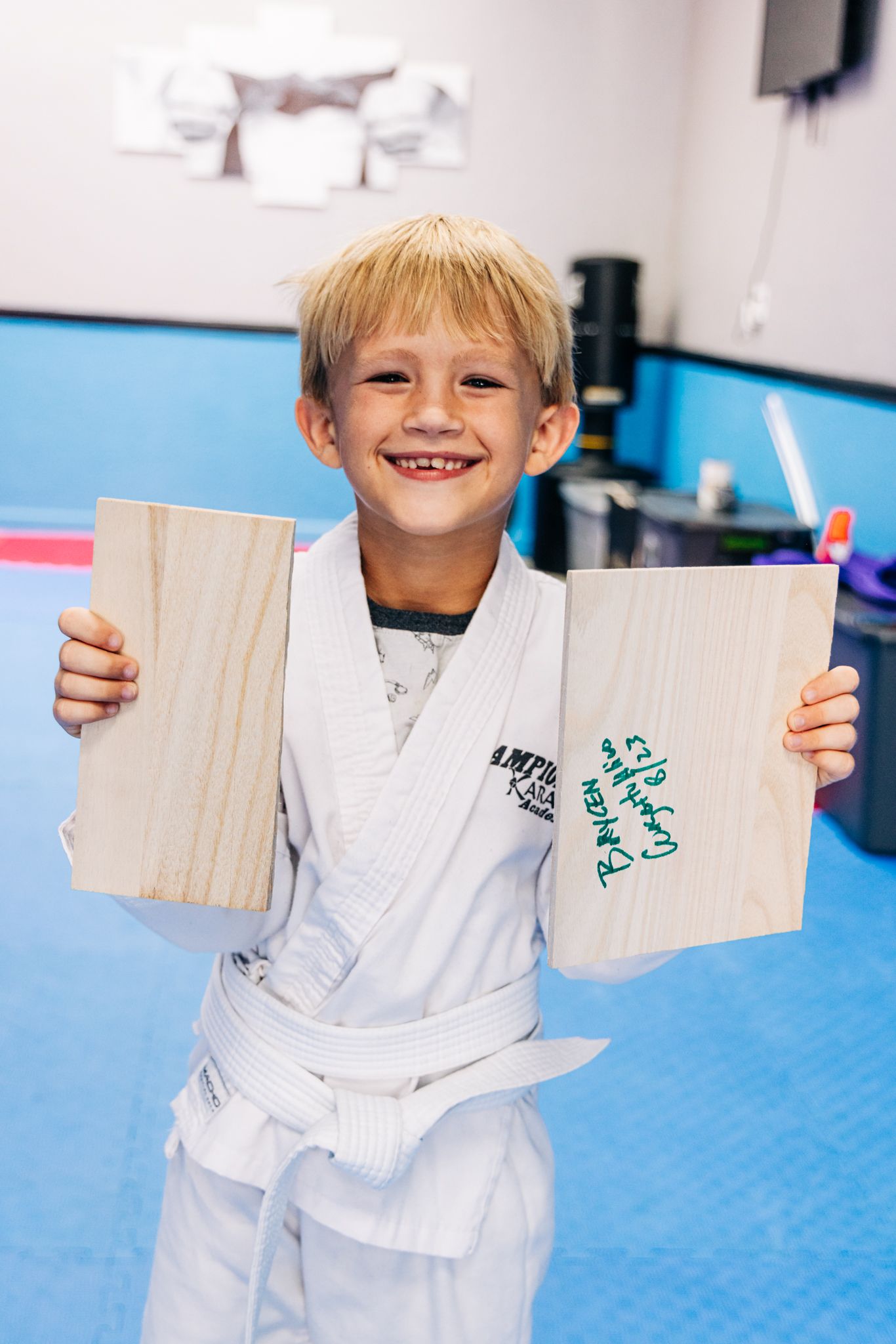 Smiling young boy in white karate gi holds two broken wooden boards after a successful board-breaking achievement in martial arts class.