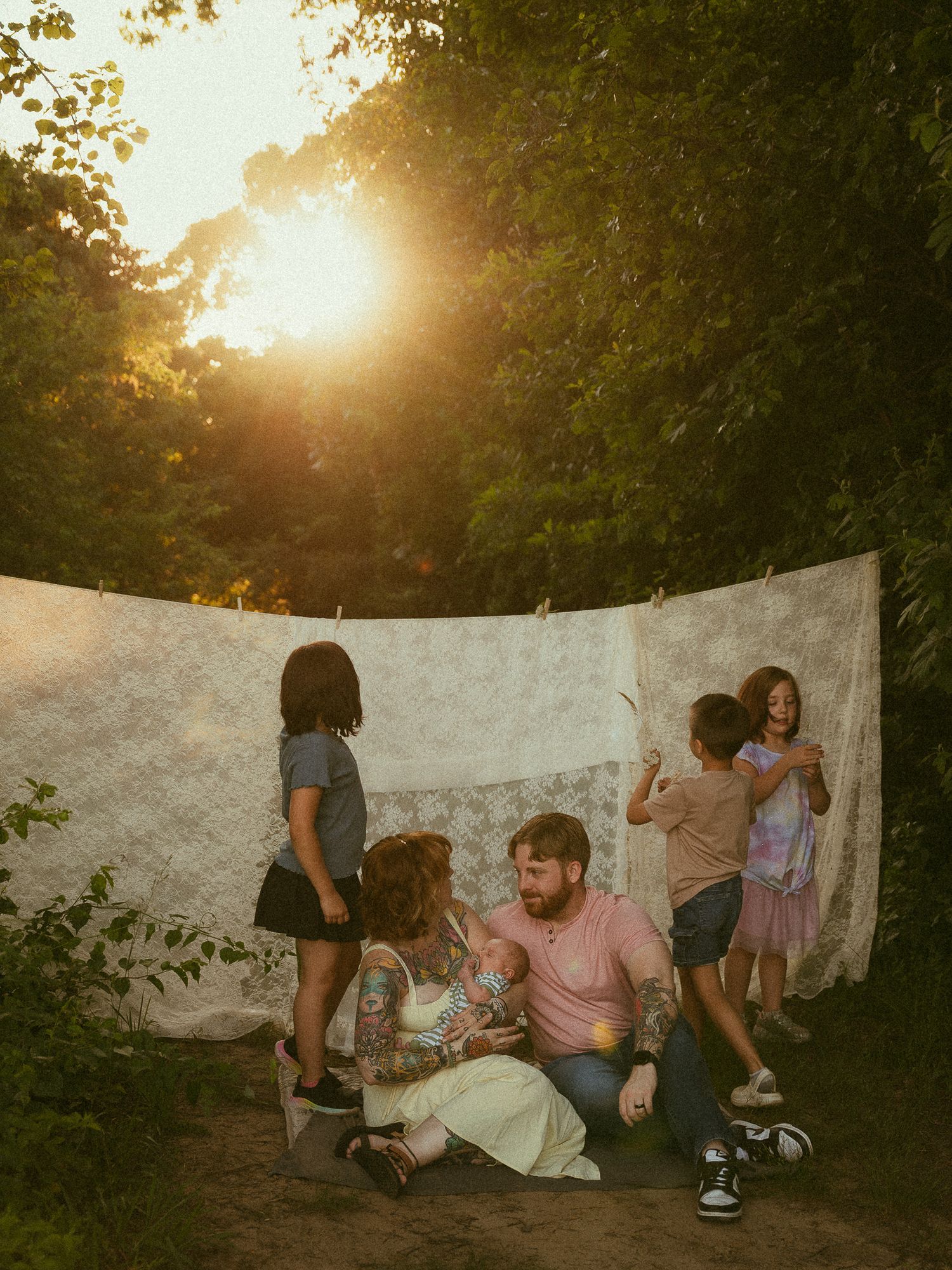 Family sitting together outdoors at golden hour near a hanging lace cloth, surrounded by children playing in a wooded setting.