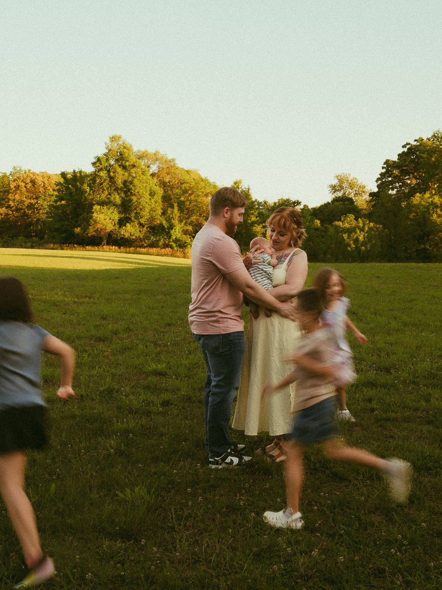 Family with baby stands in sunlit green field while children run around them at golden hour.