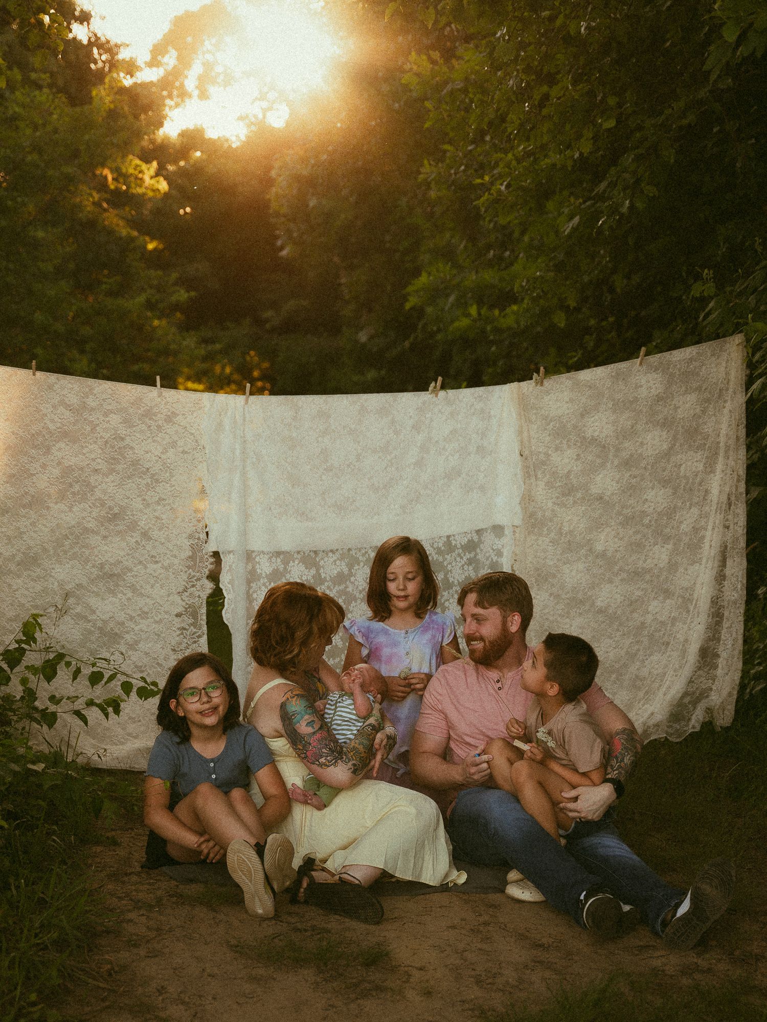 Family of six sitting together on ground in front of lace sheets on clothesline at golden hour sunset outdoors.