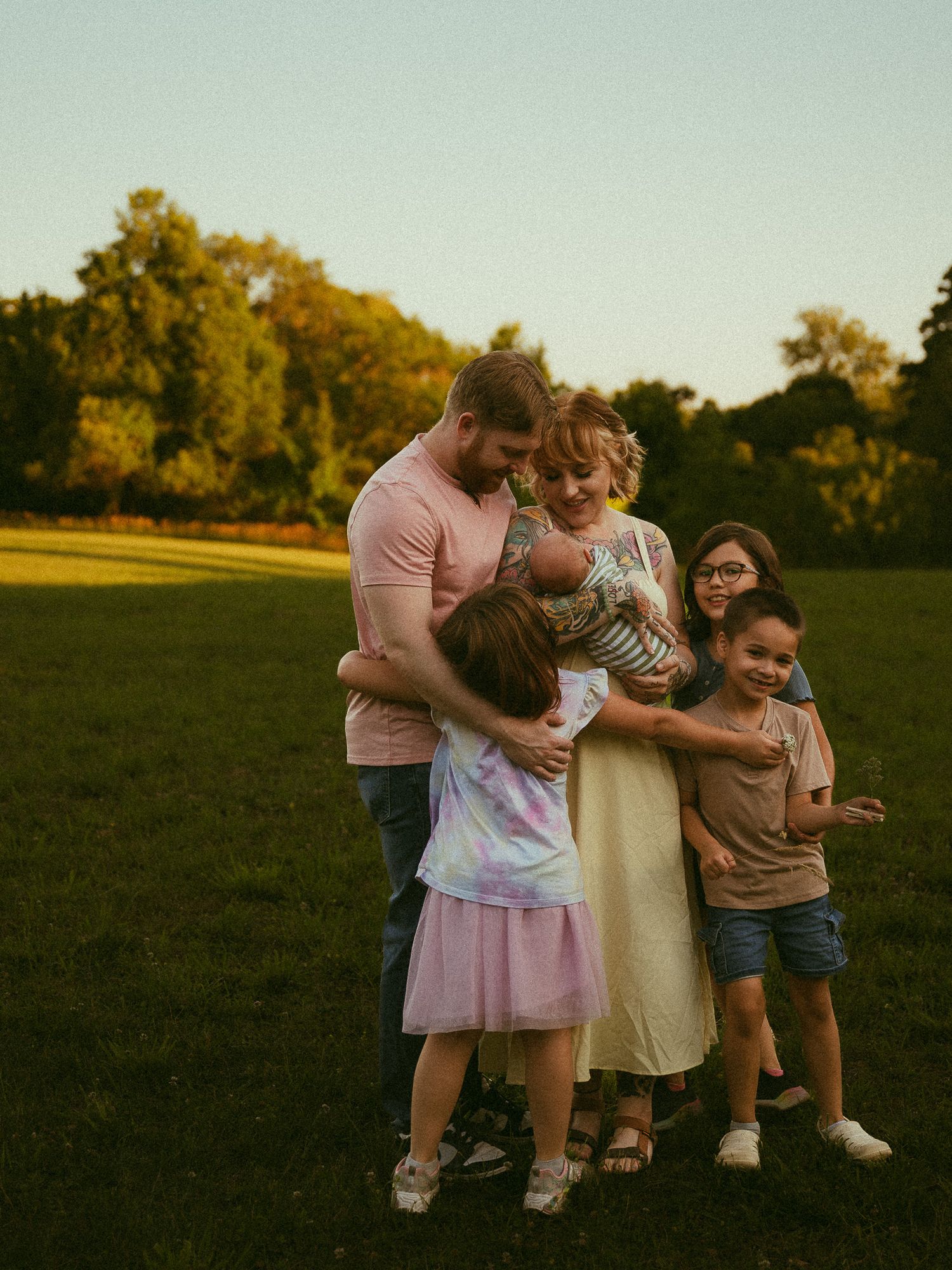 Family of six sharing a warm embrace in a sunlit green field during golden hour, with trees in the background.