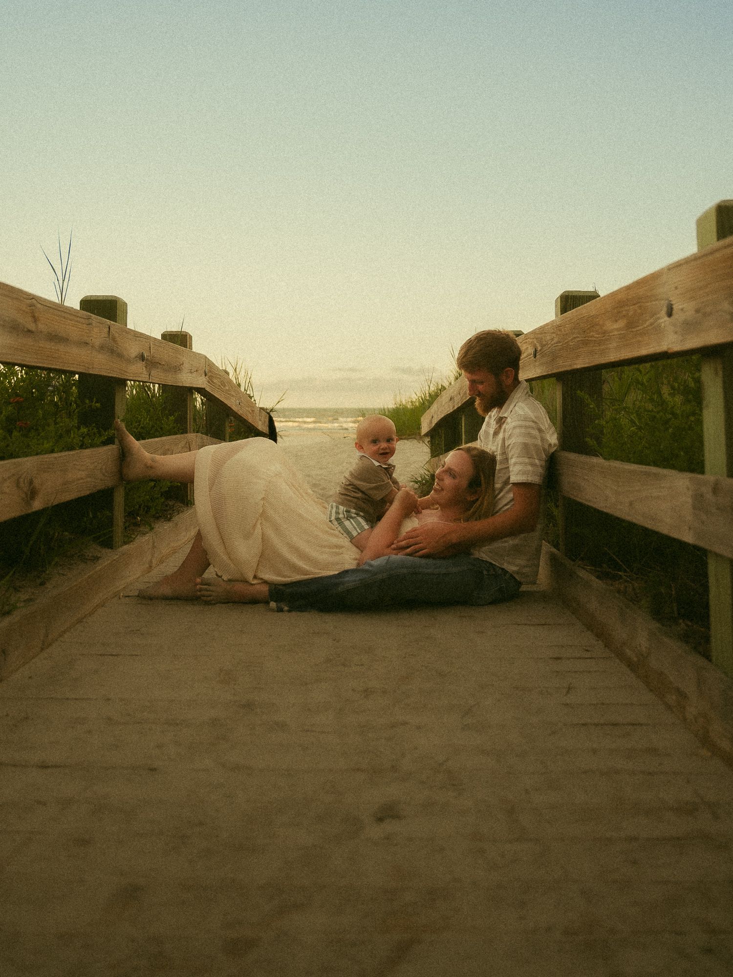 Family sitting on a wooden boardwalk near the beach at sunset, with parents and a baby sharing a tender moment.