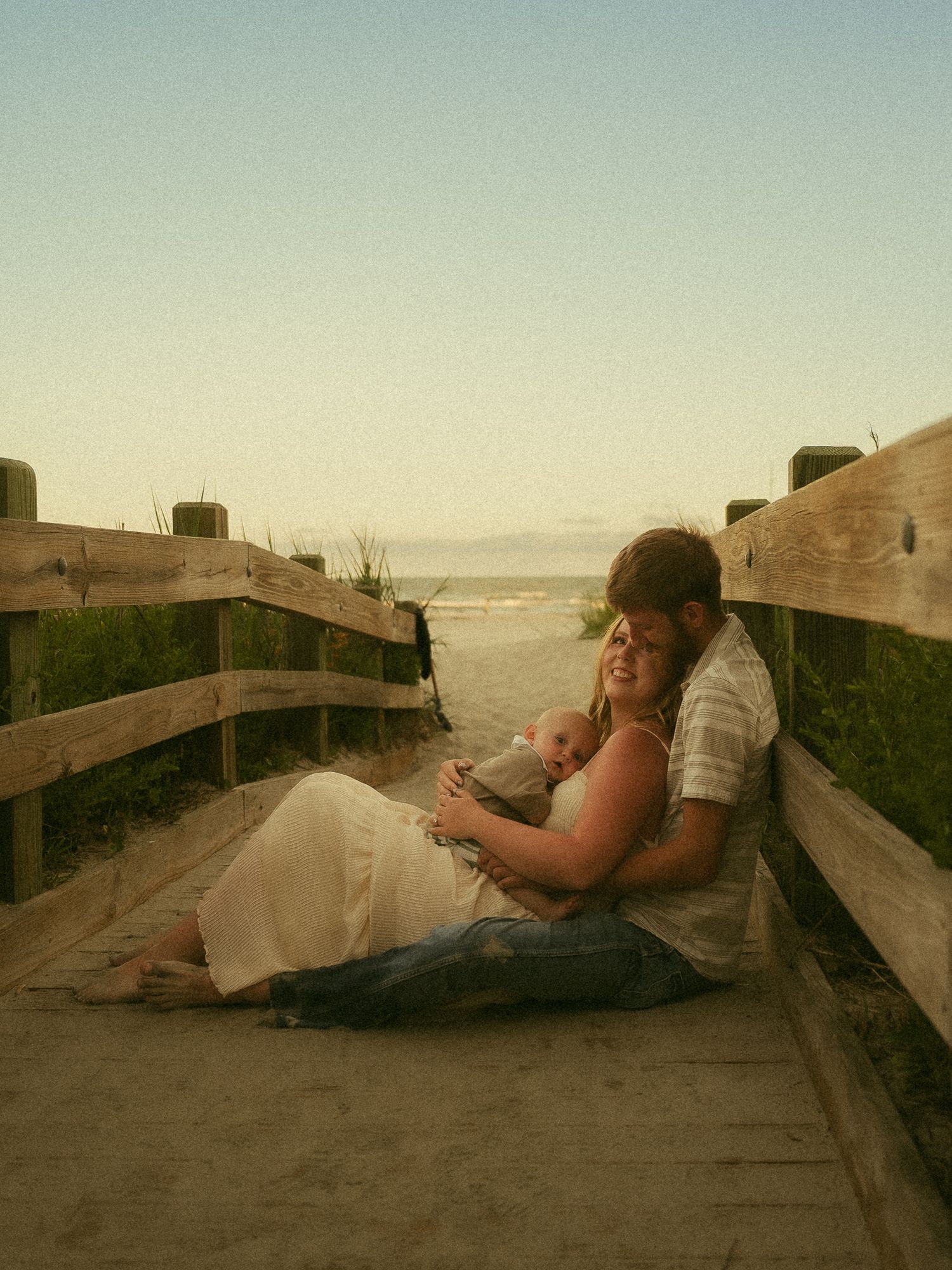 Couple sitting on a wooden boardwalk at the beach, embracing with a baby at sunset.