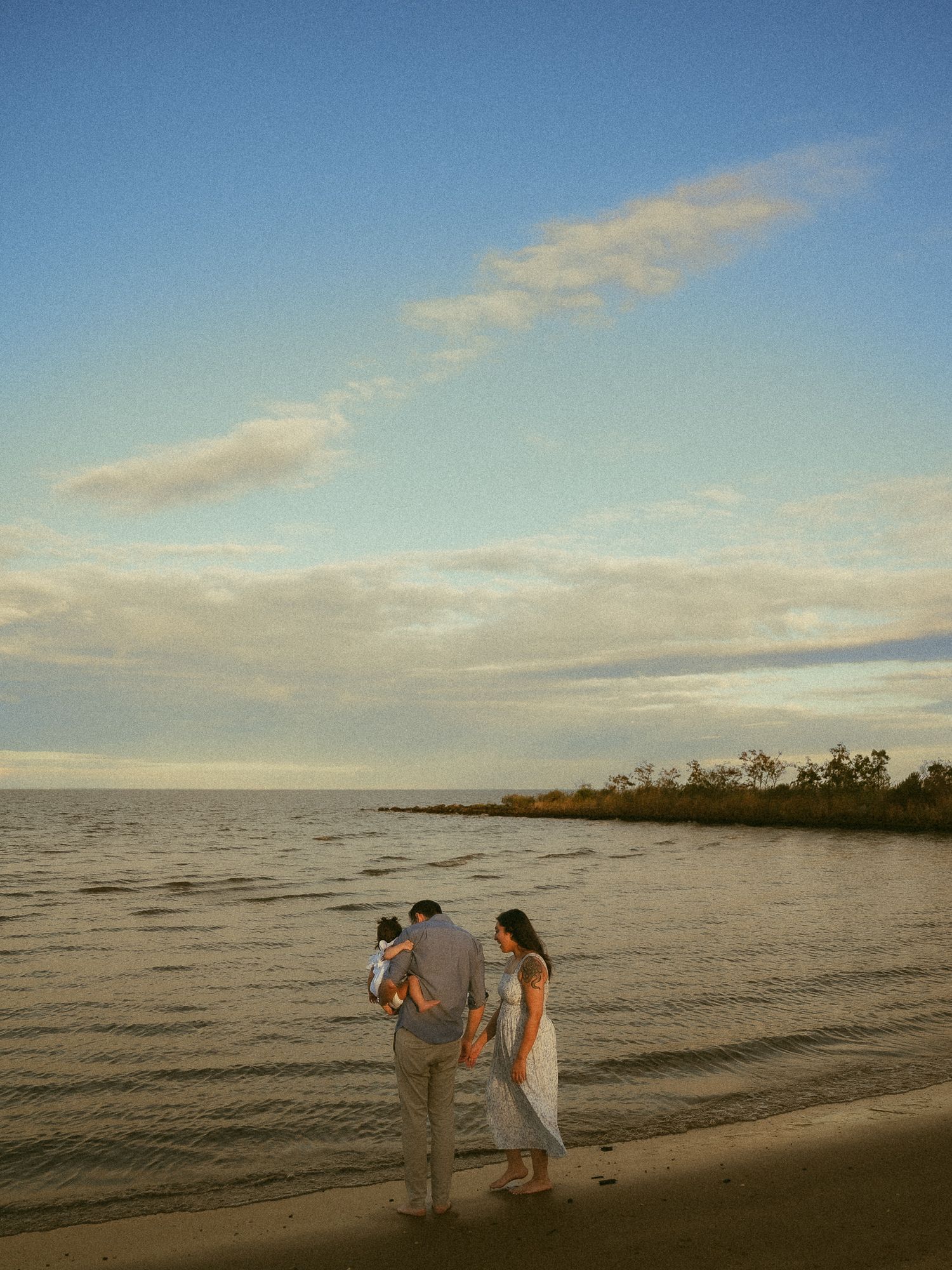 Family of three standing on a sandy beach at golden hour, gazing at calm water under a pastel blue sky.