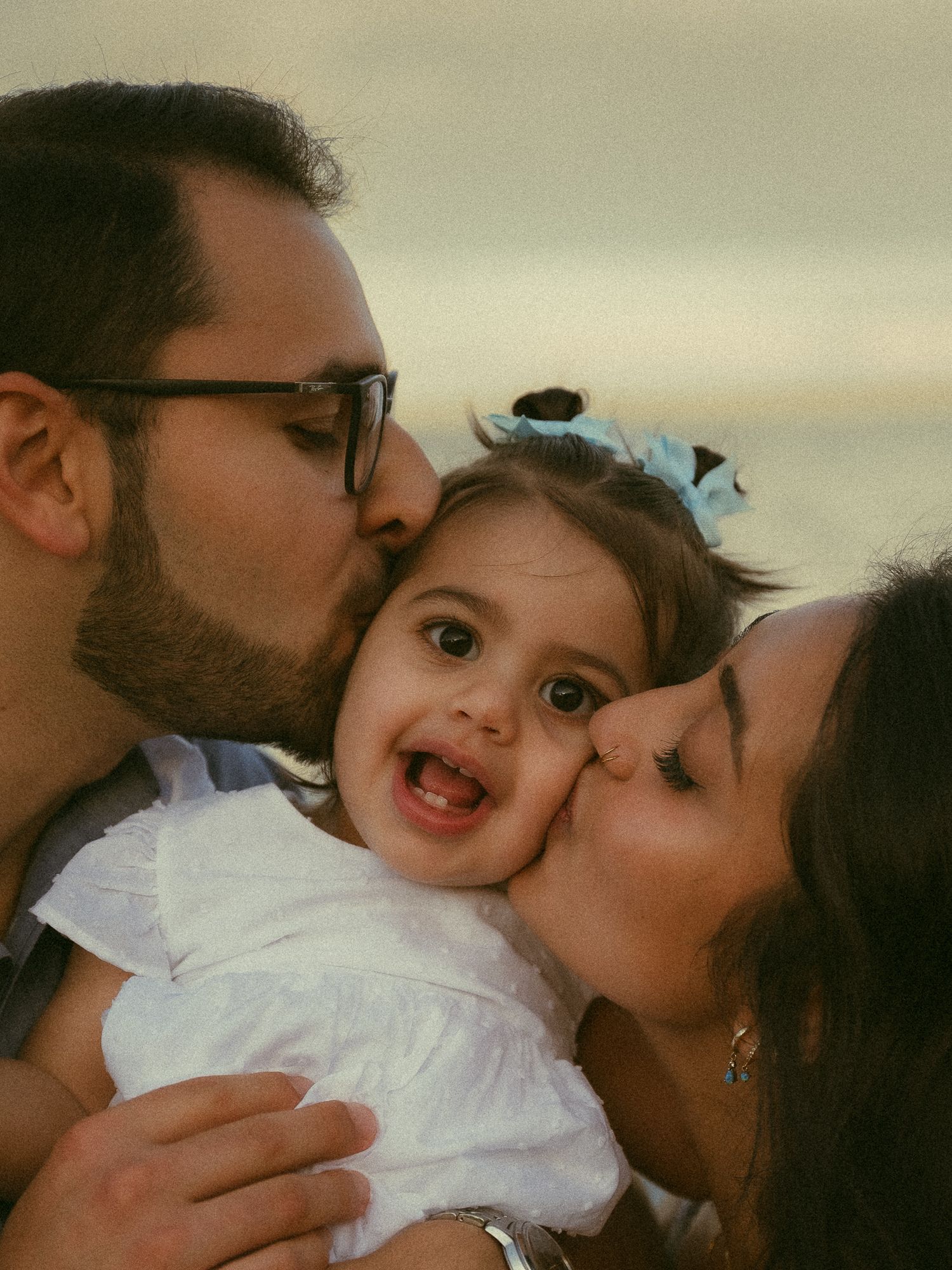 Two parents lovingly kiss their smiling baby girl wearing a white dress and blue bow on a beach.
