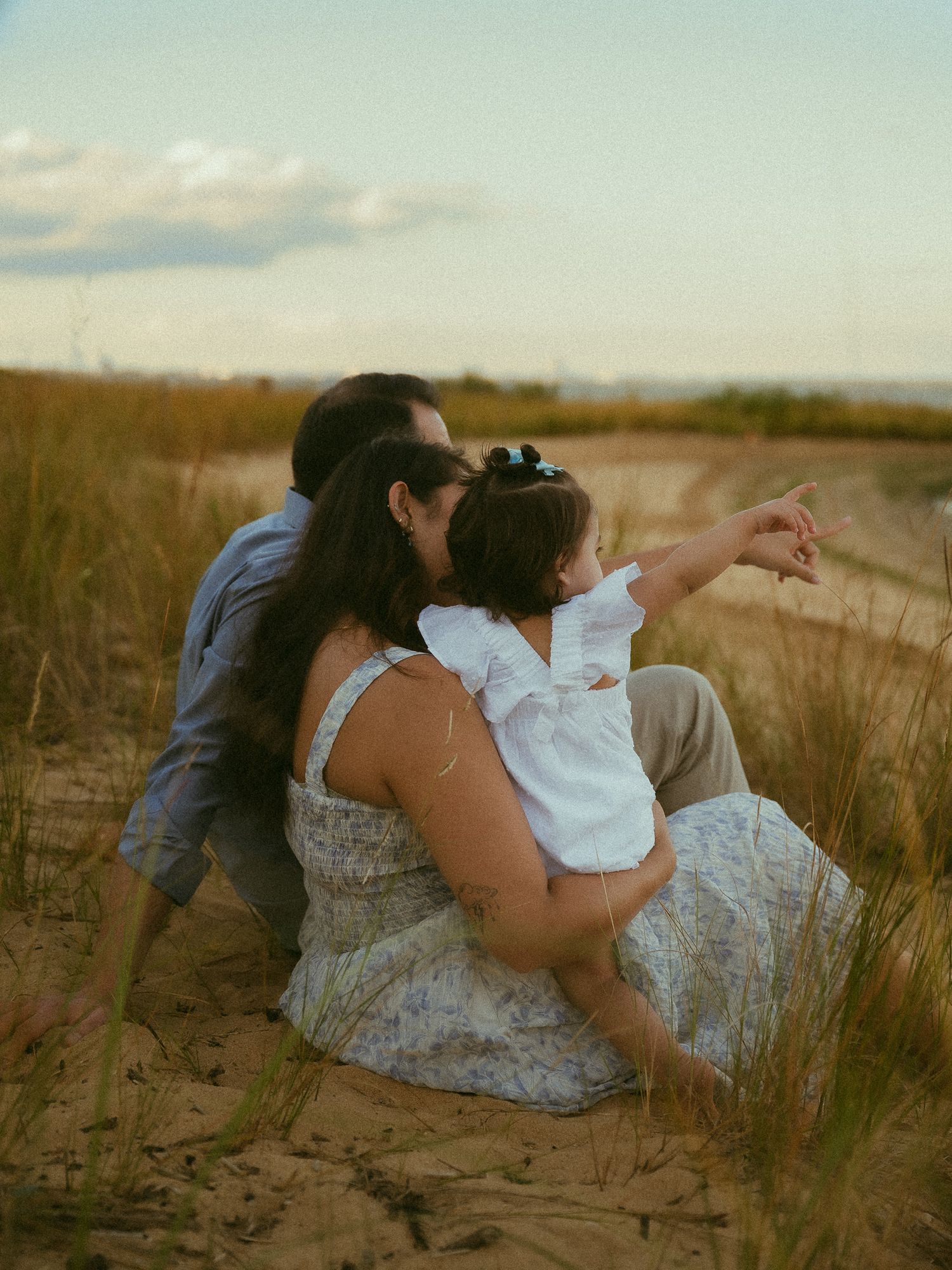 A couple sits on sandy dunes at sunset, cradling a toddler in white who points toward the horizon.