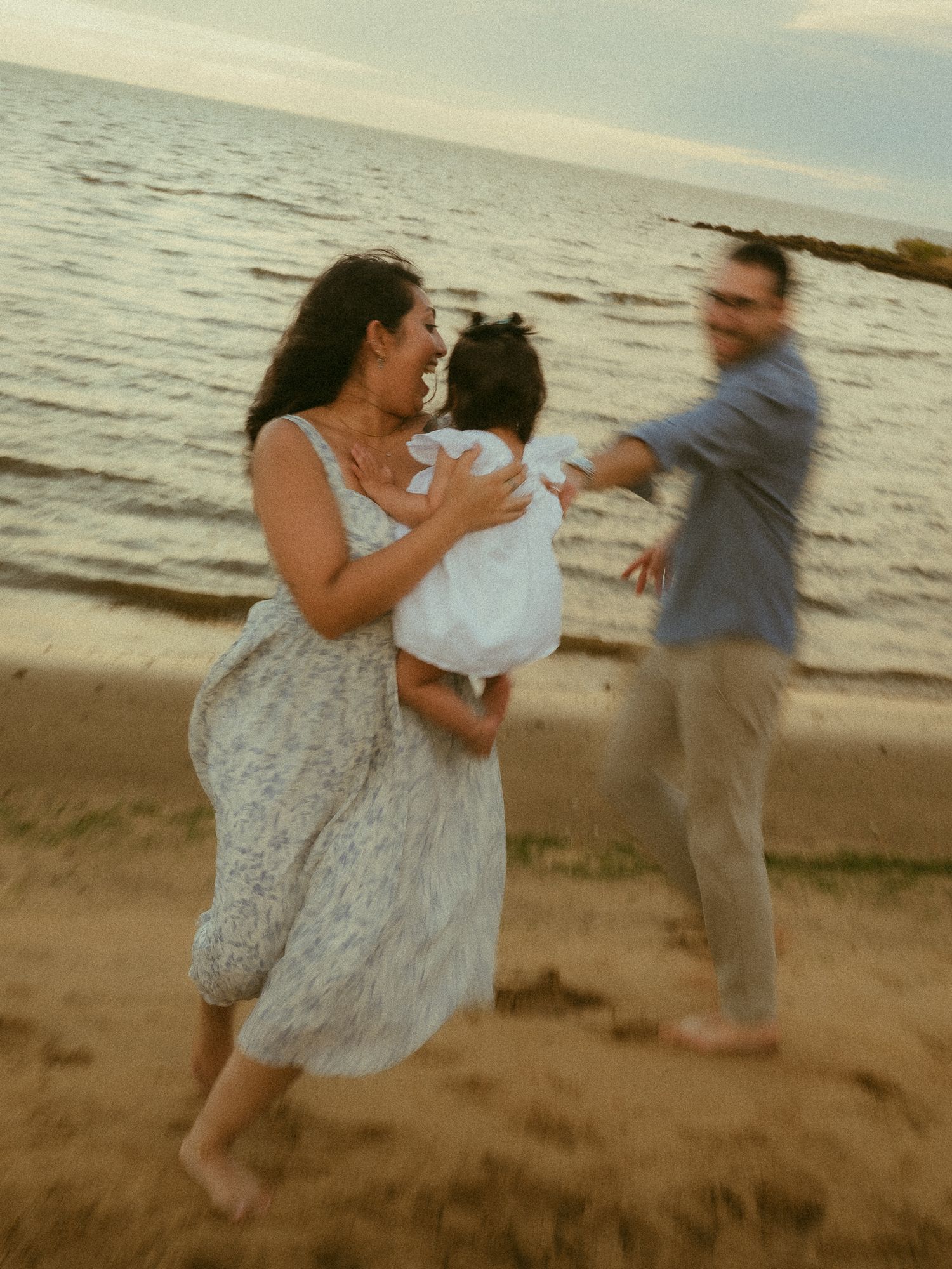 A joyful family plays on a sandy beach at sunset, with a woman in a floral dress holding a baby in white.