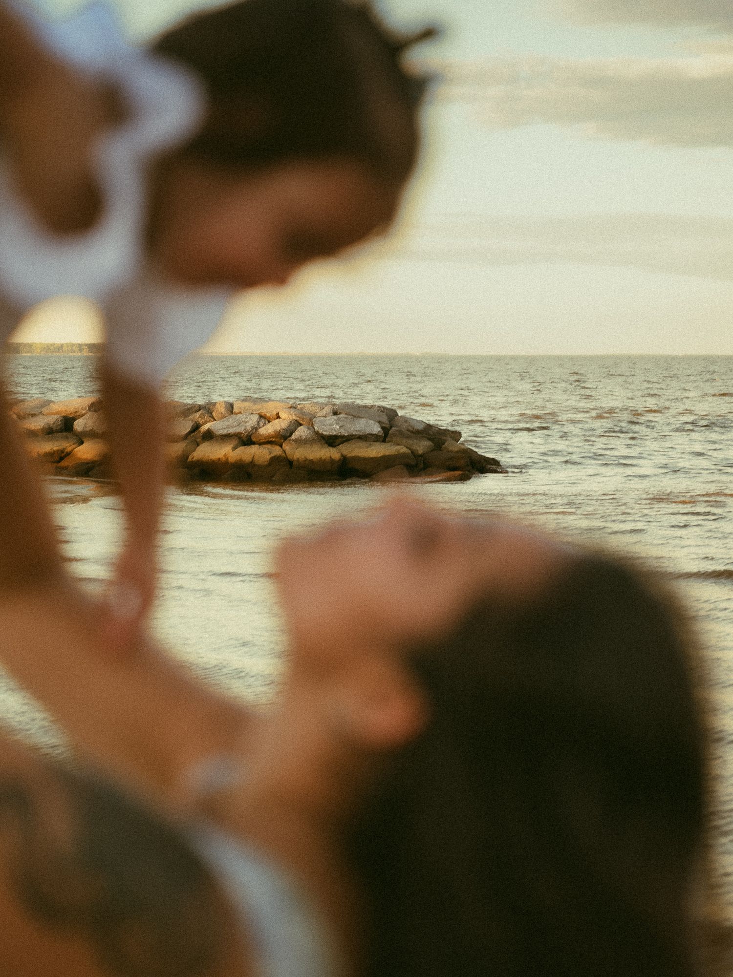 A mother lifts her baby at a serene beach during golden hour, with rocky formations visible in the calm ocean.
