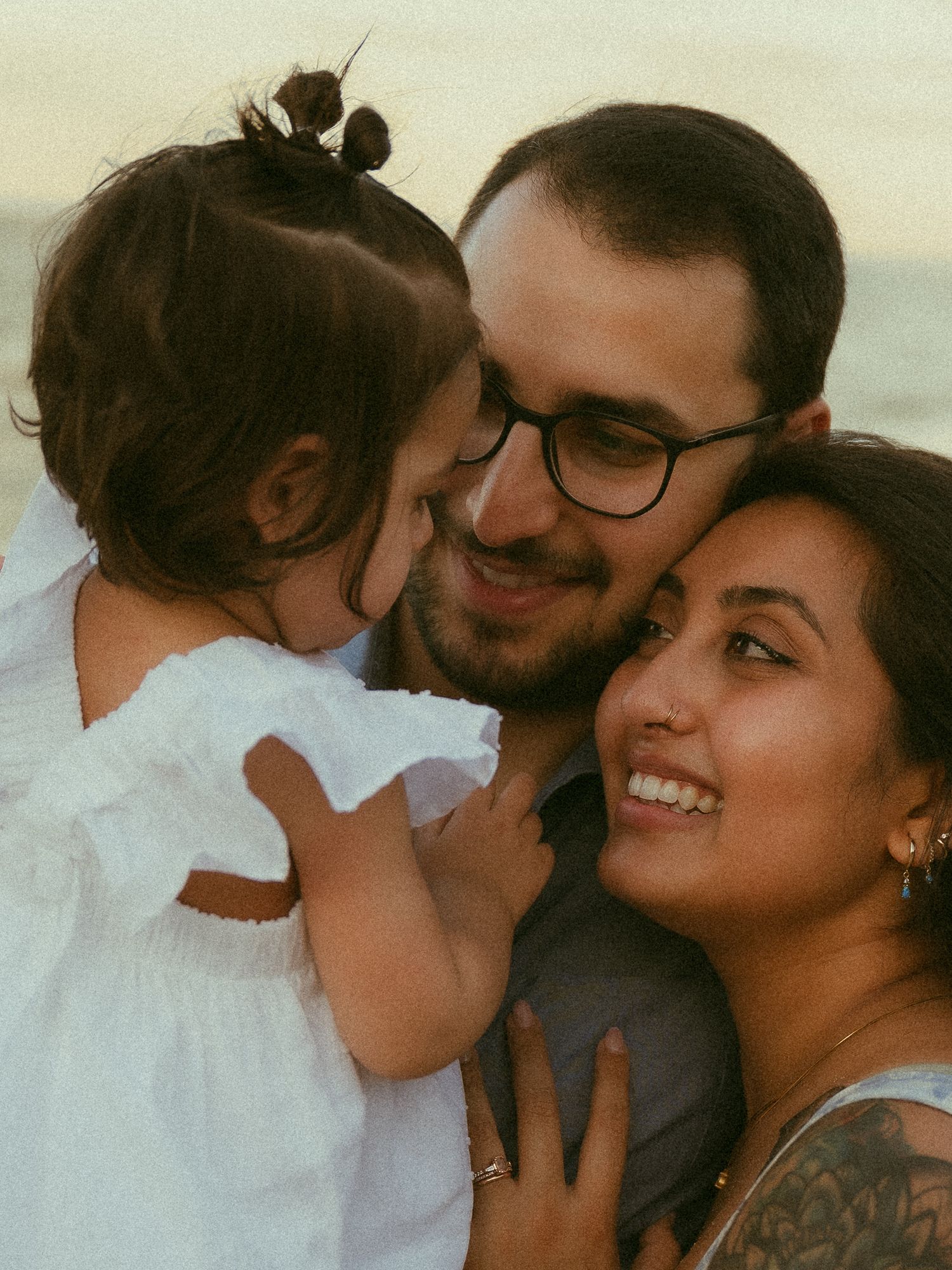 A happy family of three sharing a tender moment, with parents smiling at their toddler daughter in a white dress.