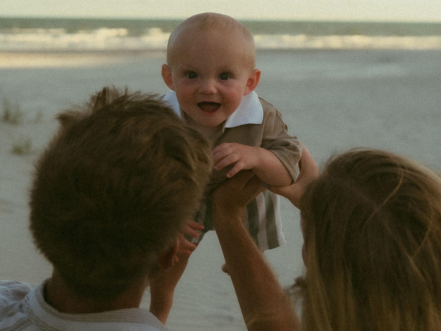 Parents lifting smiling baby at beach during sunset, family moment on sandy shore with ocean waves in background.