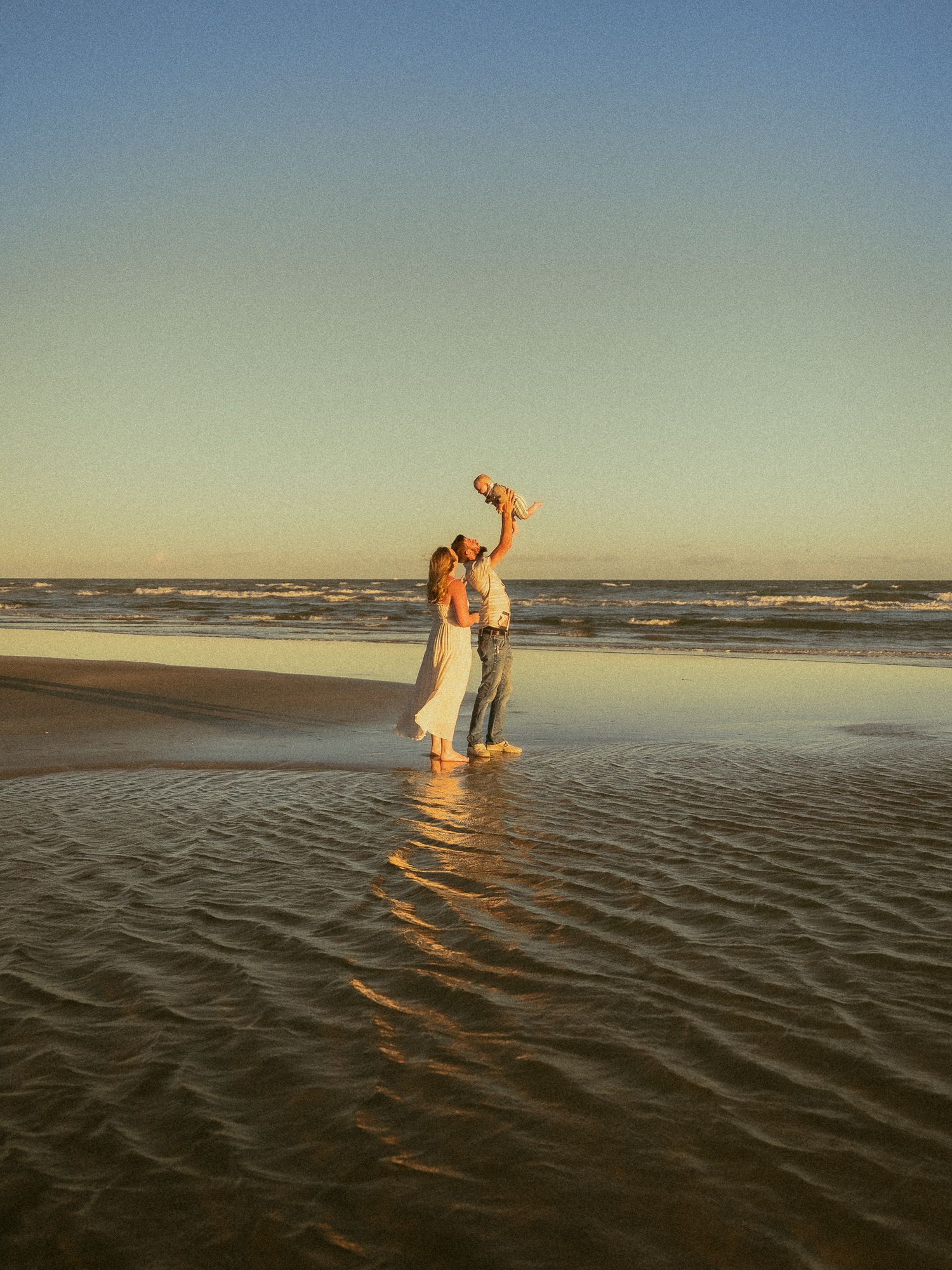 Couple lifting baby at sunset on a reflective beach with ocean waves in the background.