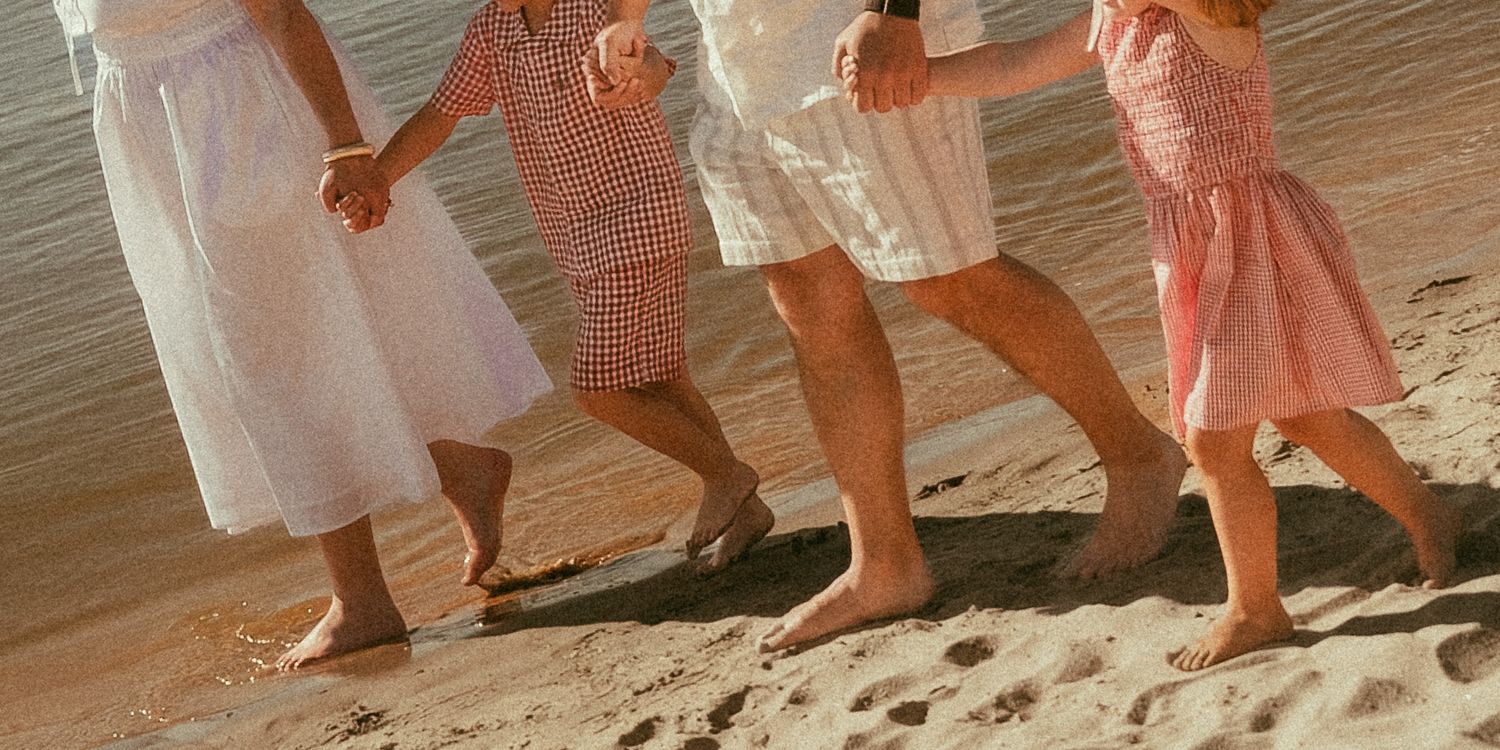 Four women in summer dresses walk barefoot together on a sandy beach, holding hands in the warm sunlight.