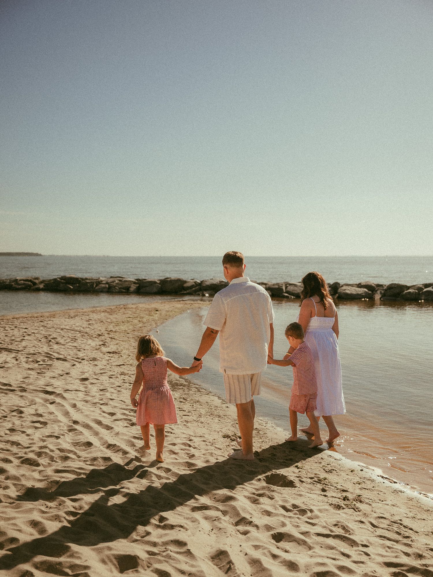 Family of four holding hands on a sandy beach at golden hour, facing the ocean in matching pink and white outfits.