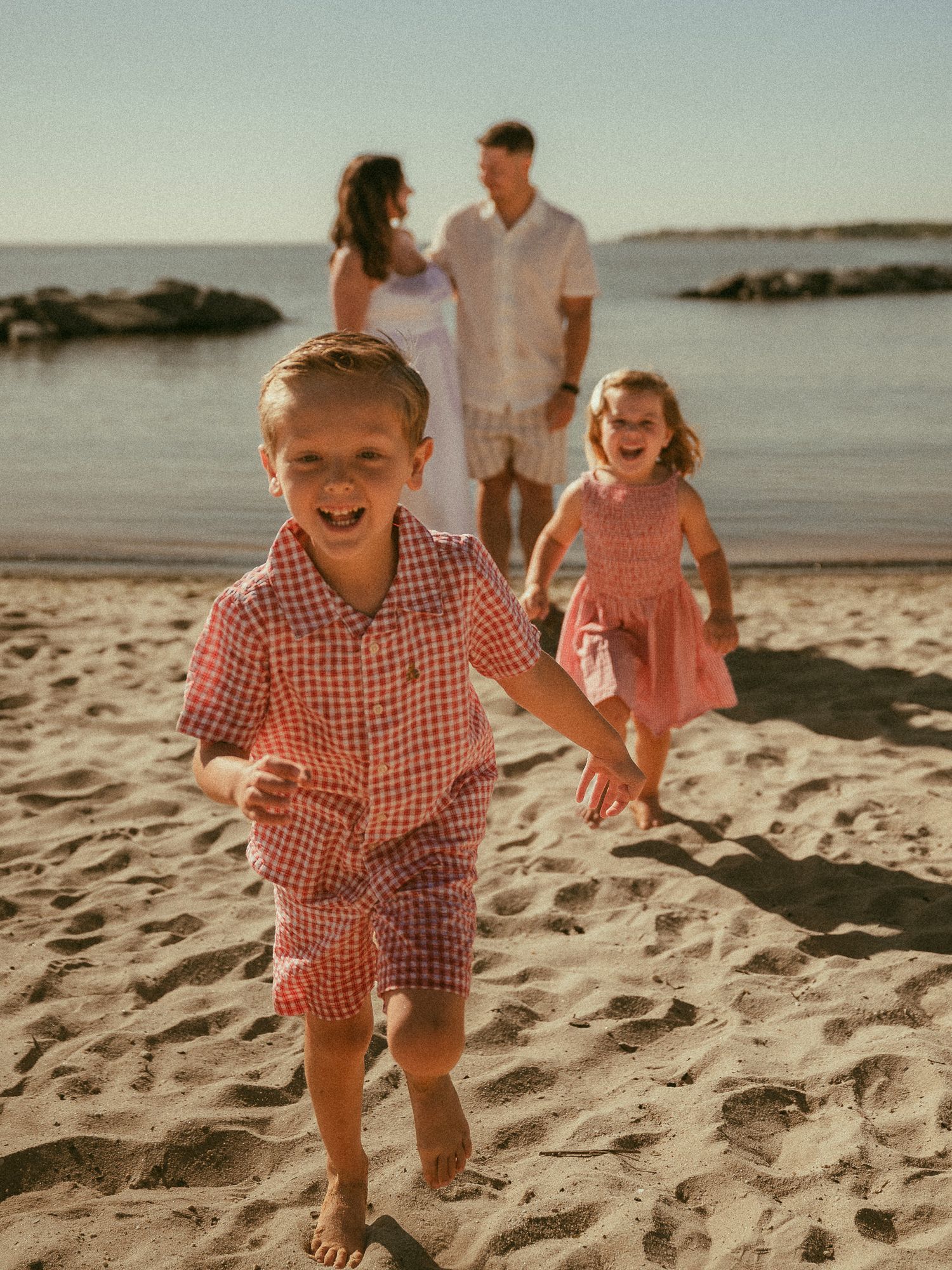 Two joyful children run on a sandy beach while their parents stand together near the water in warm golden light.