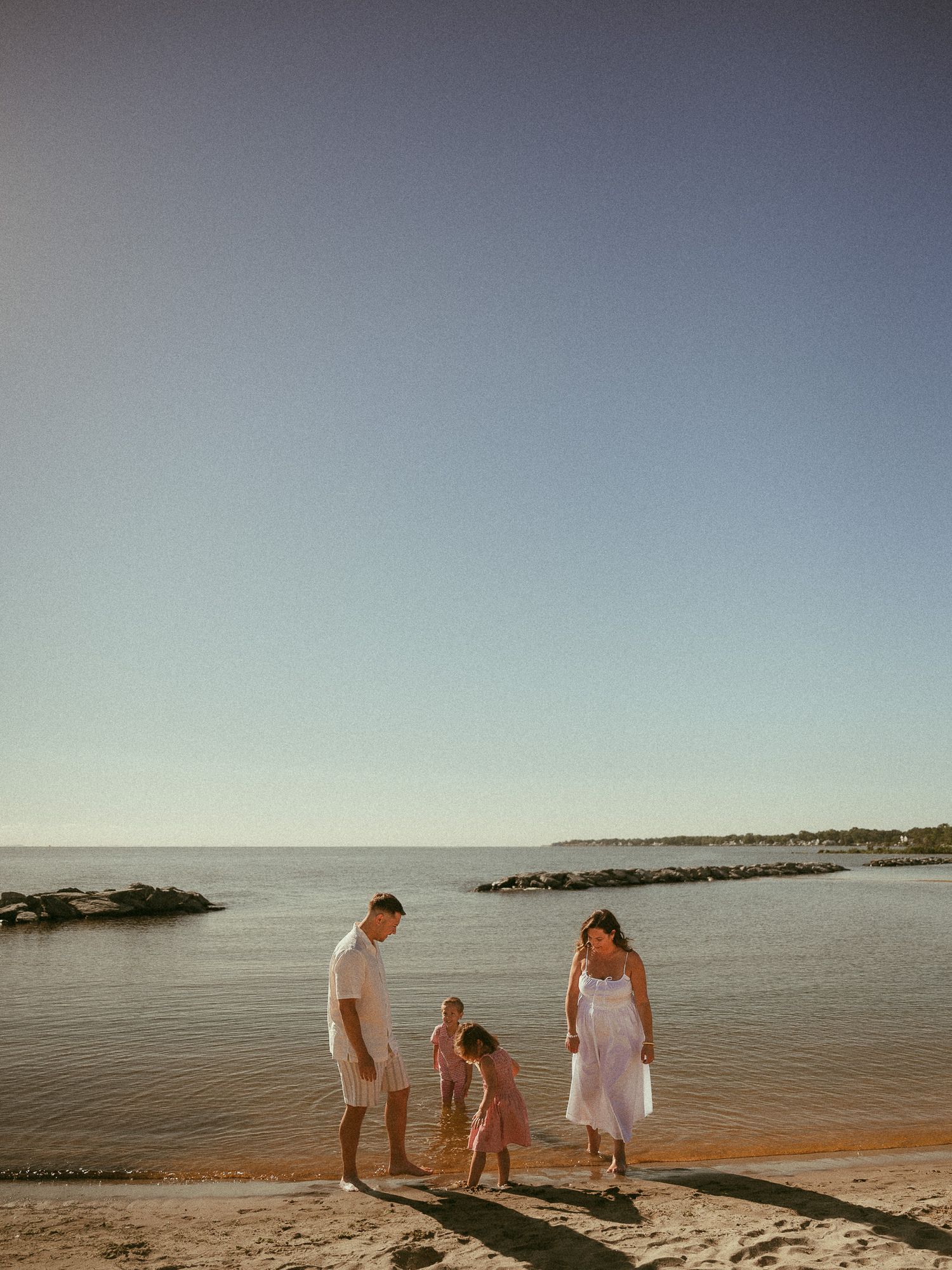 Family of three enjoying a sunny beach day, parents watching their young child play on sandy shore near calm ocean waters.
