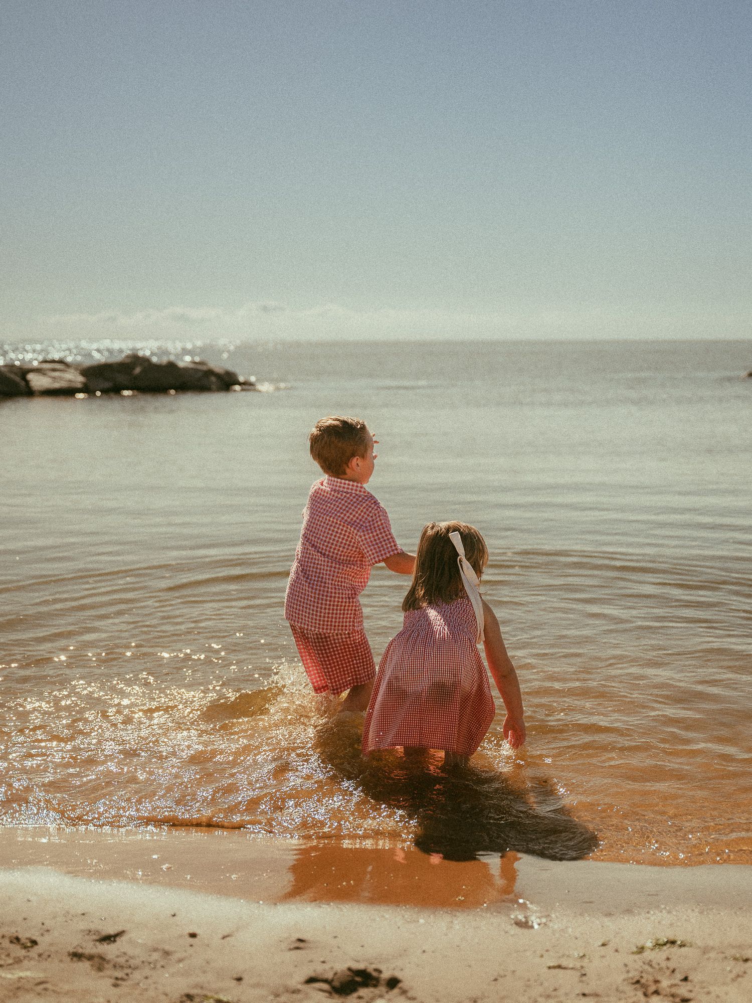 Two children in matching pink outfits wade in shallow ocean waves on a sunny beach.