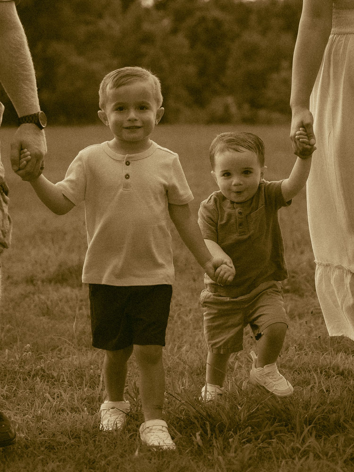 Two young boys holding hands with adults in a sepia-toned outdoor photo on a grassy field.