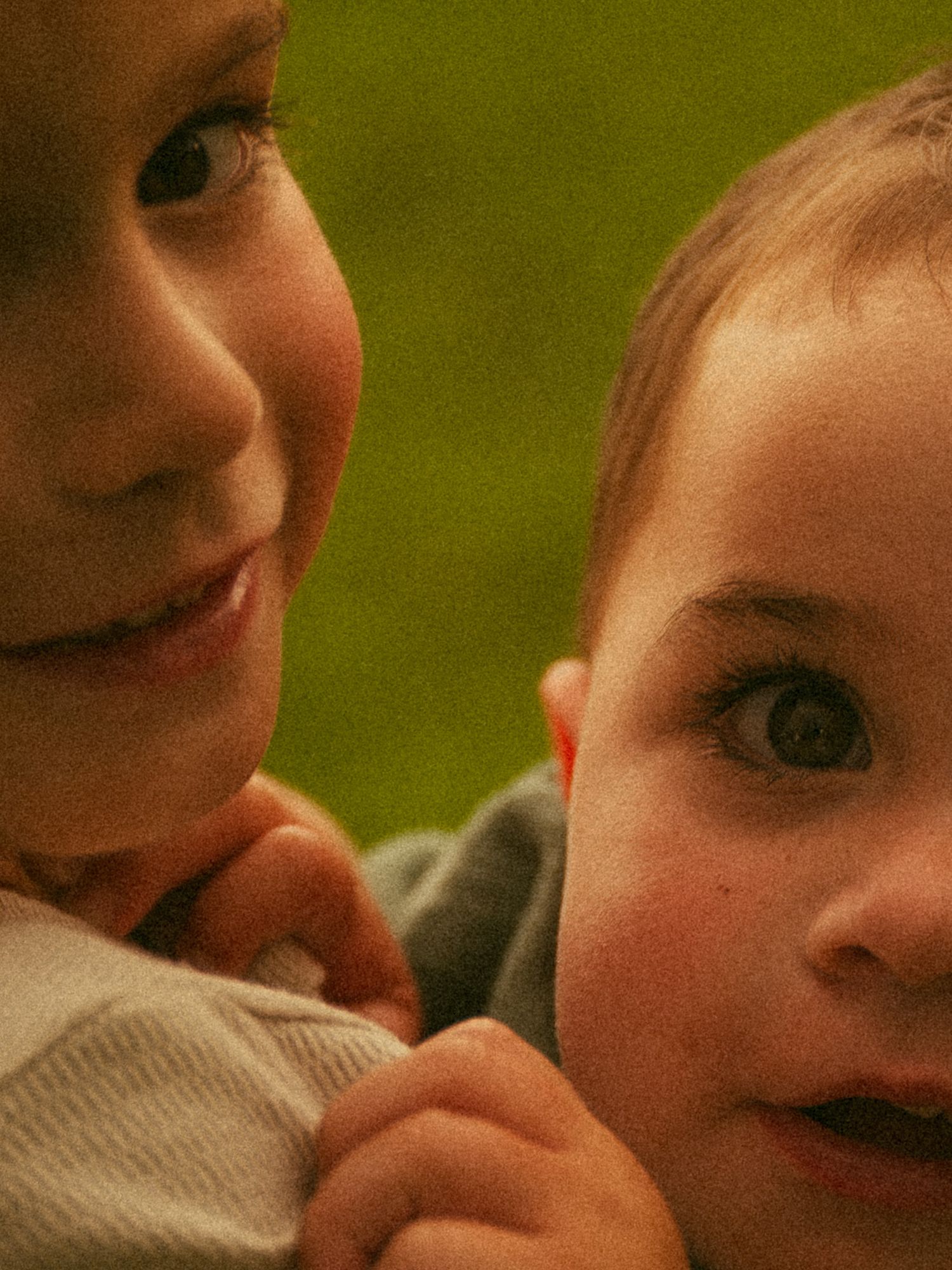 Two young children with big eyes smile outdoors, with a green blurred background, capturing a sweet sibling moment.