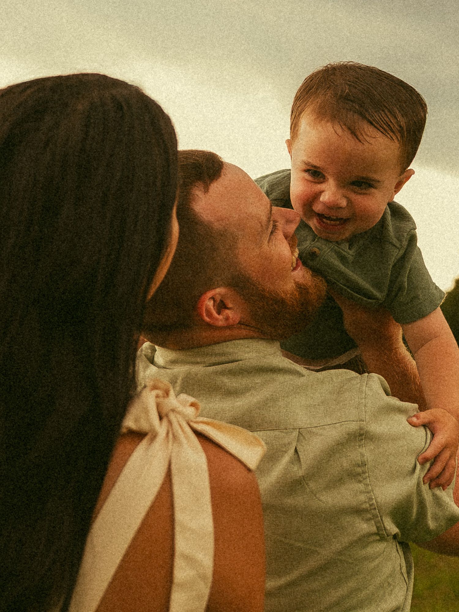 A bearded father lifts his laughing baby boy while a dark-haired woman stands beside them outdoors.