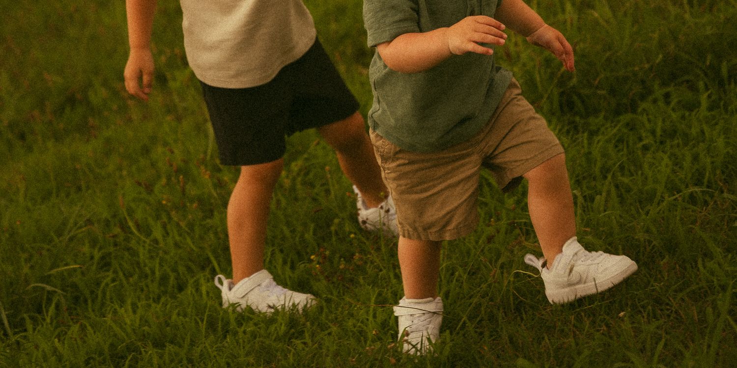 Two children running and playing on lush green grass, wearing white sneakers, shorts, and casual tops.