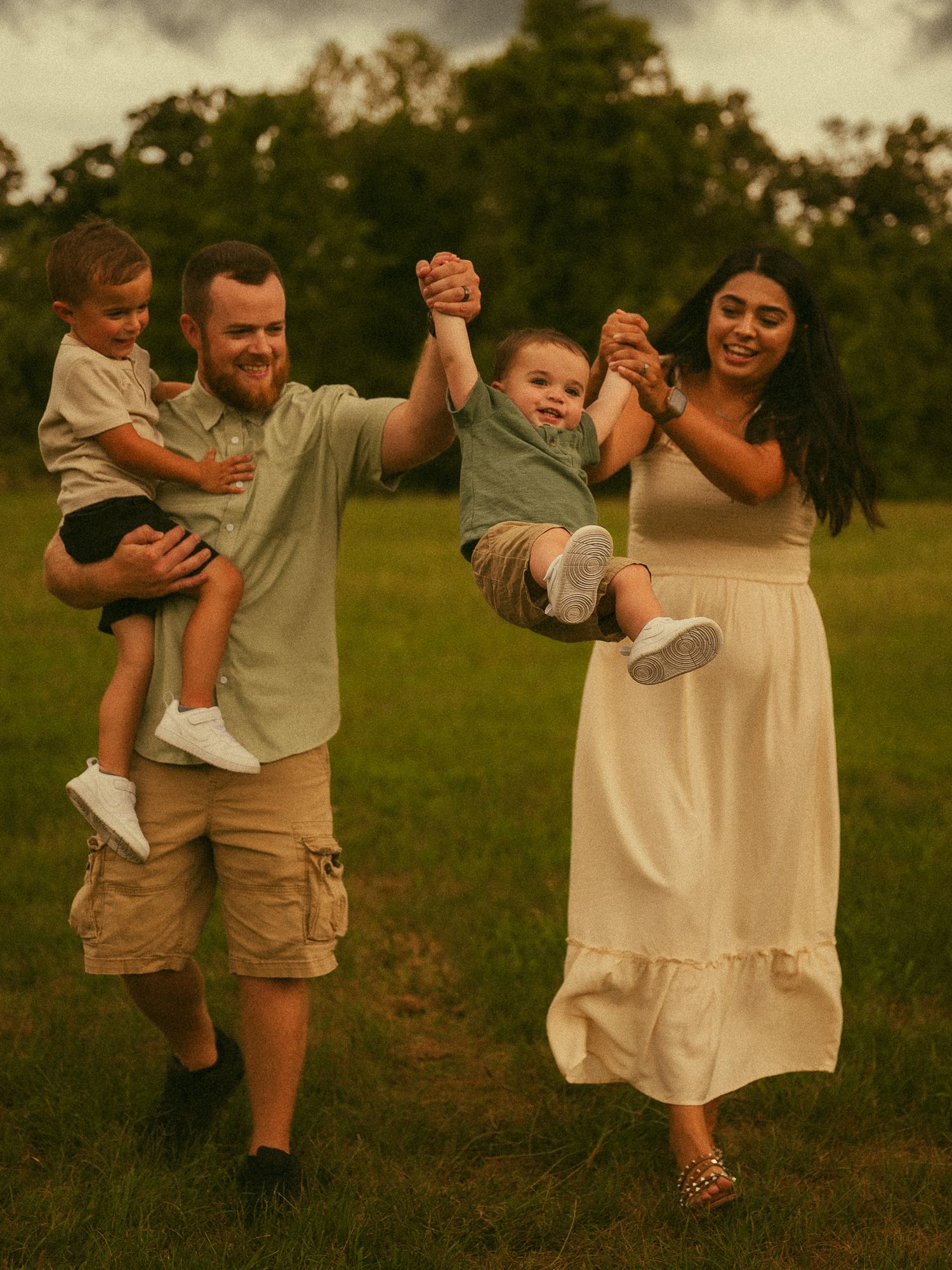 A happy family of four plays in a green field, parents swinging a toddler while holding another young child.