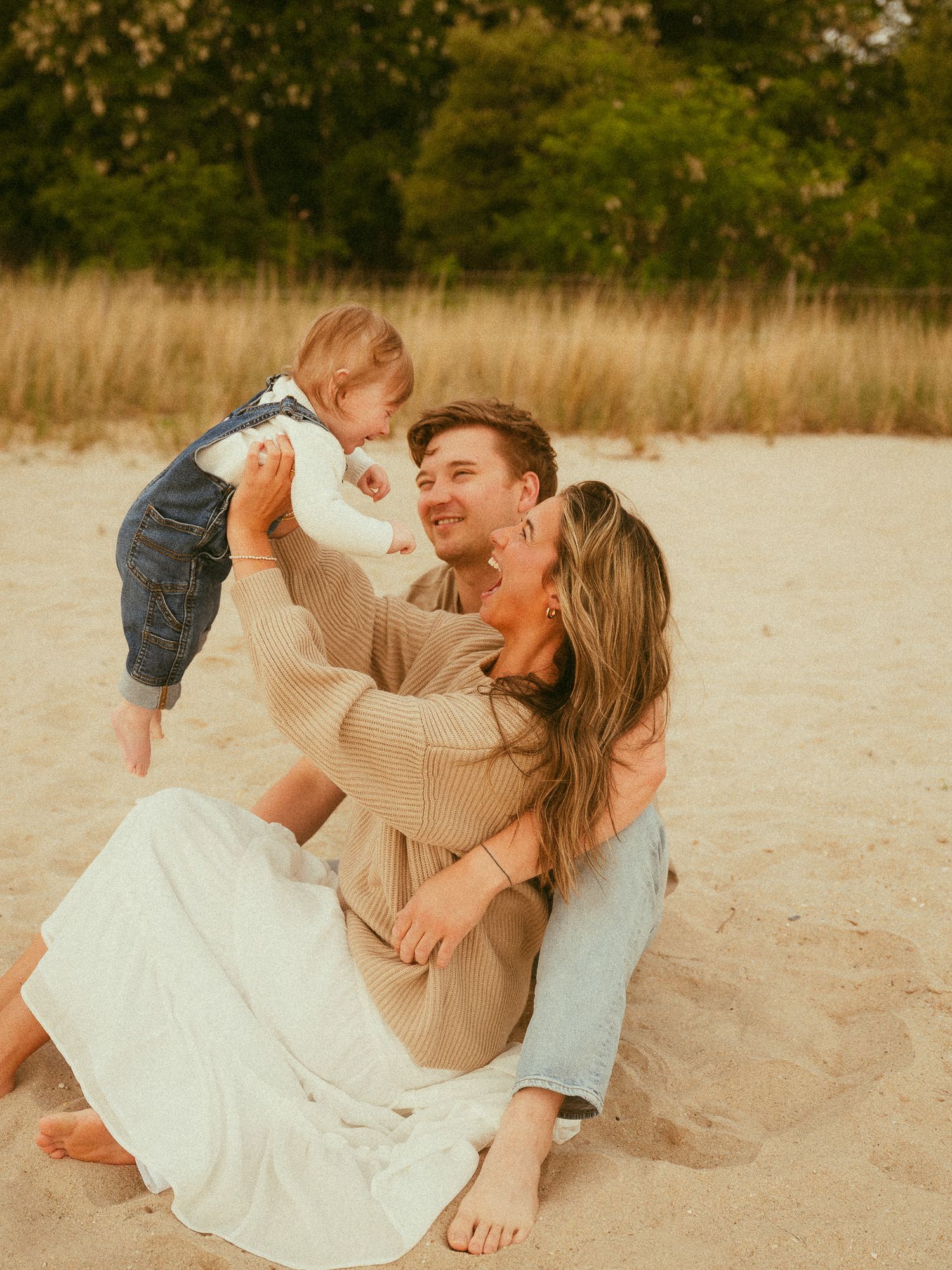 Young couple sitting on sandy beach lifting smiling baby in denim overalls with trees in background.