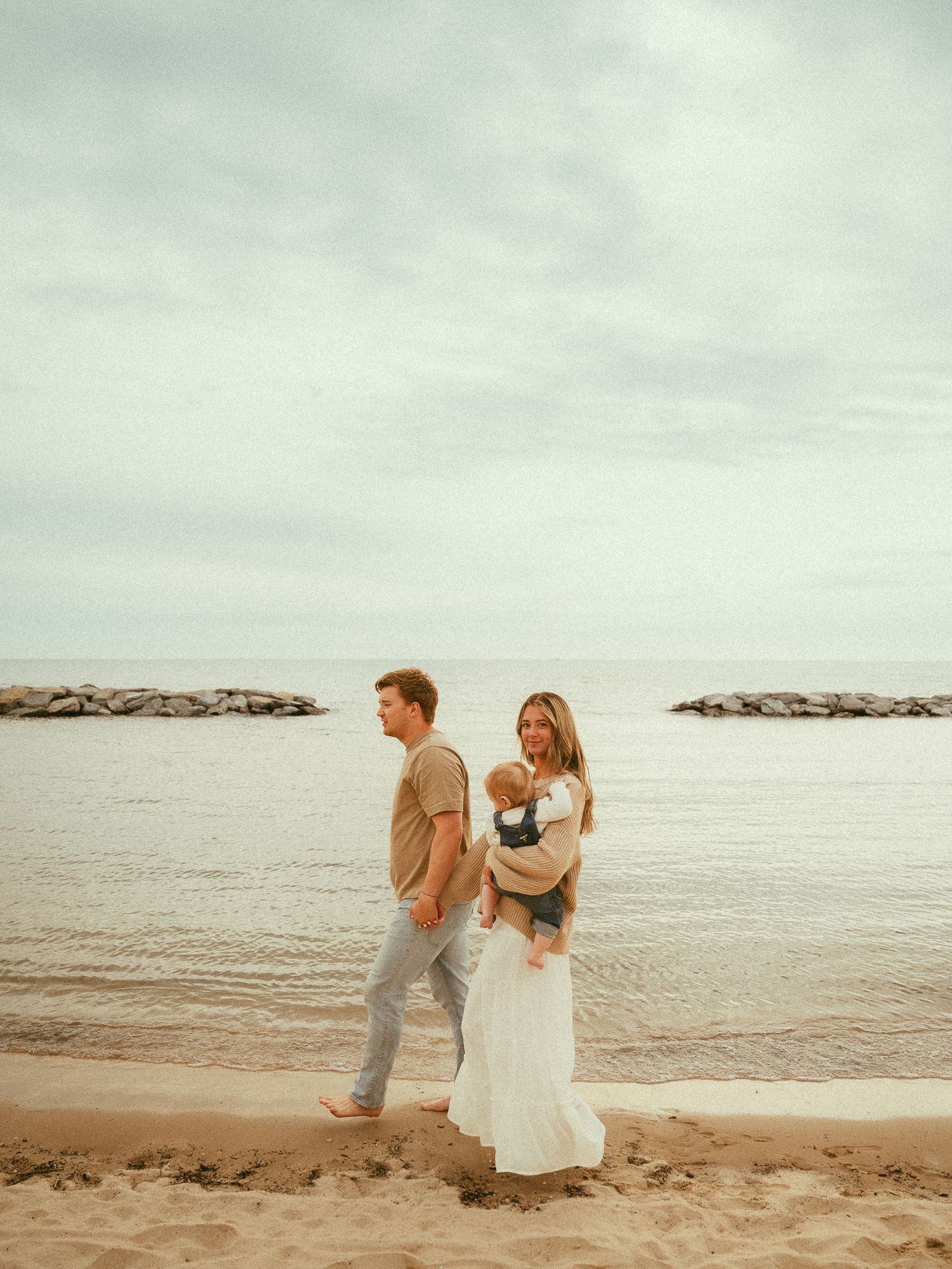 Young family walking on sandy beach shoreline, mother holding baby in carrier, wearing white dress.