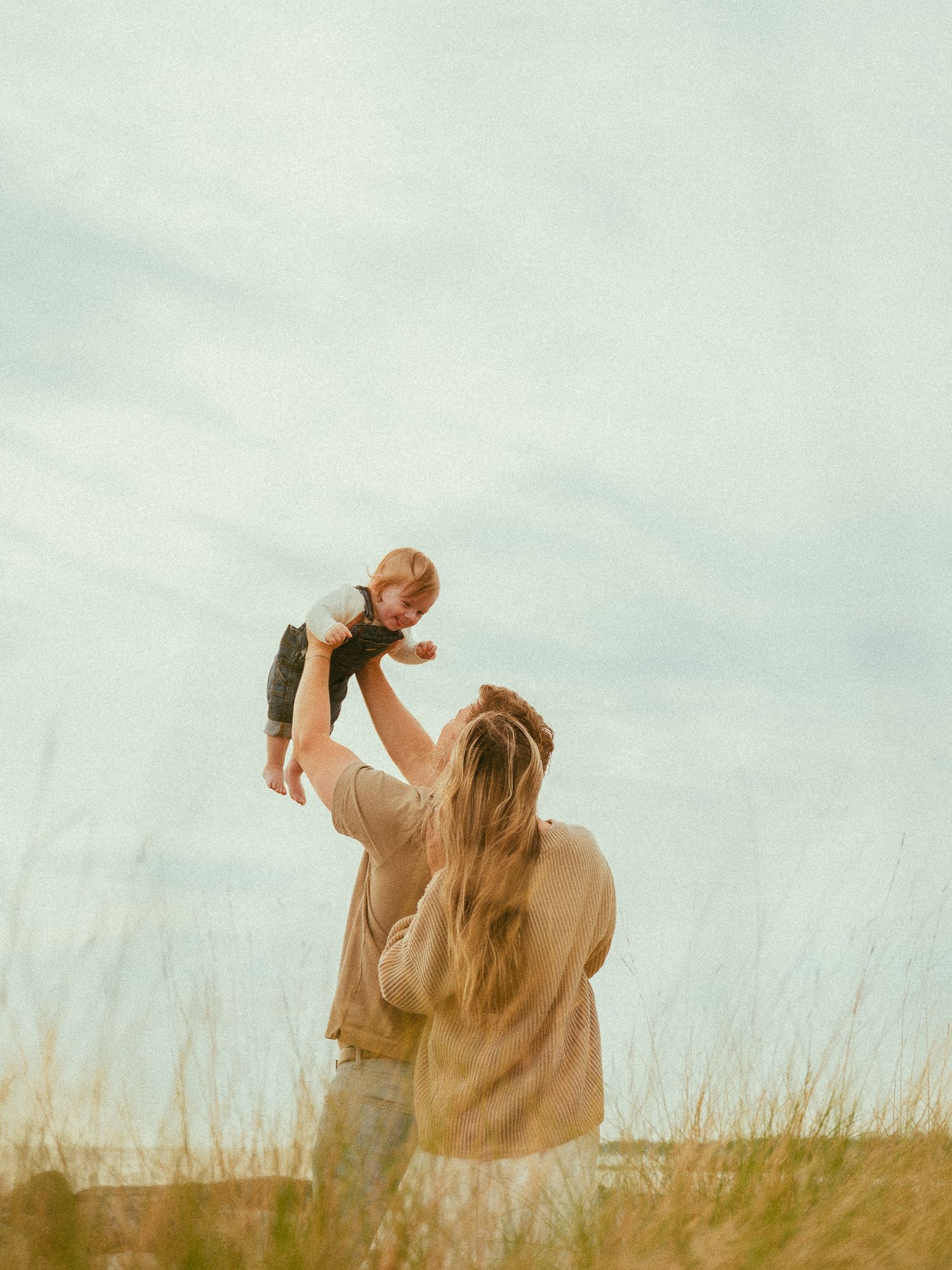 A mother lifts her baby joyfully in the air outdoors among golden grasses under a soft cloudy sky.
