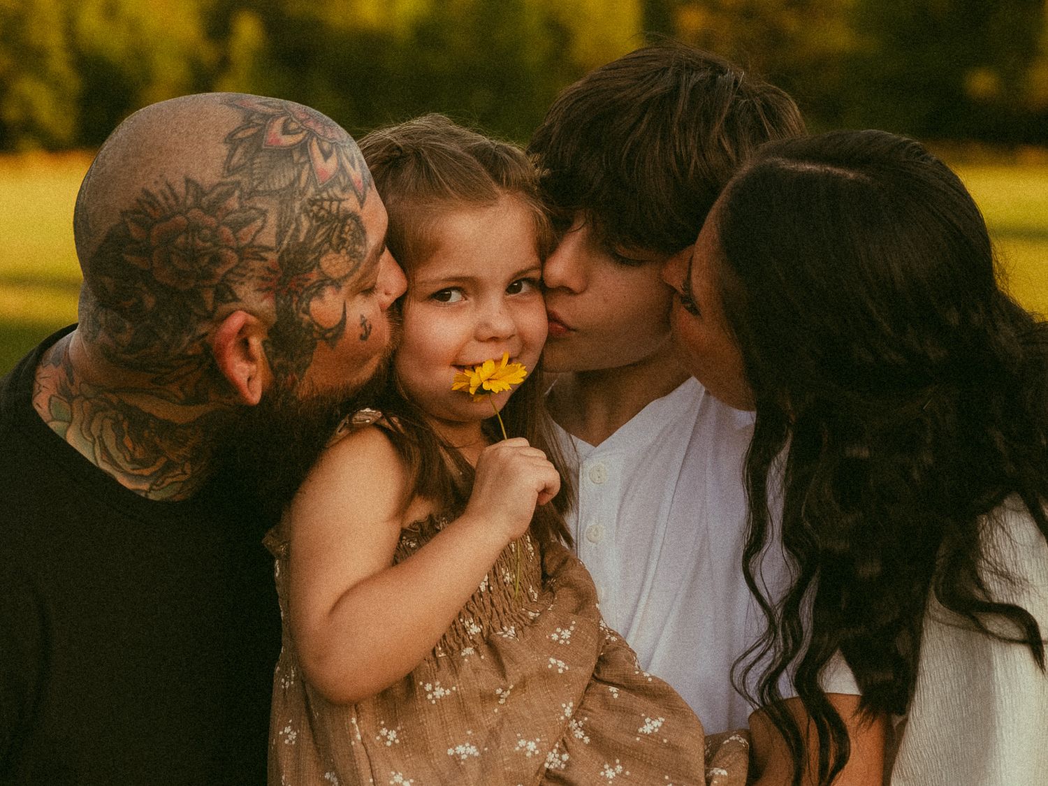 A heavily tattooed man and a woman kiss a young girl holding a yellow flower outdoors in warm golden light.