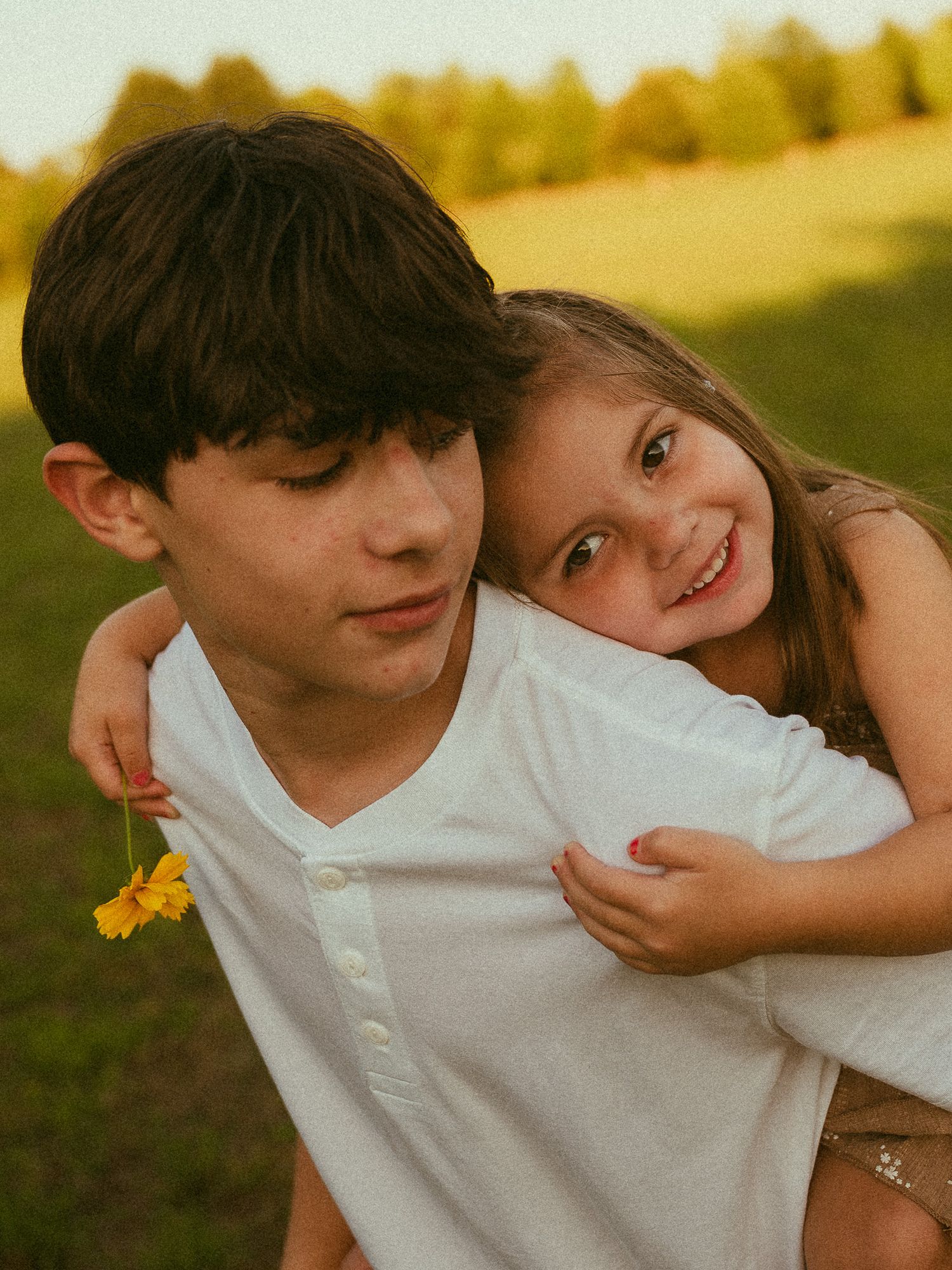 A boy in a white shirt gives a smiling girl a piggyback ride in a sunny green field, she holds a yellow flower.