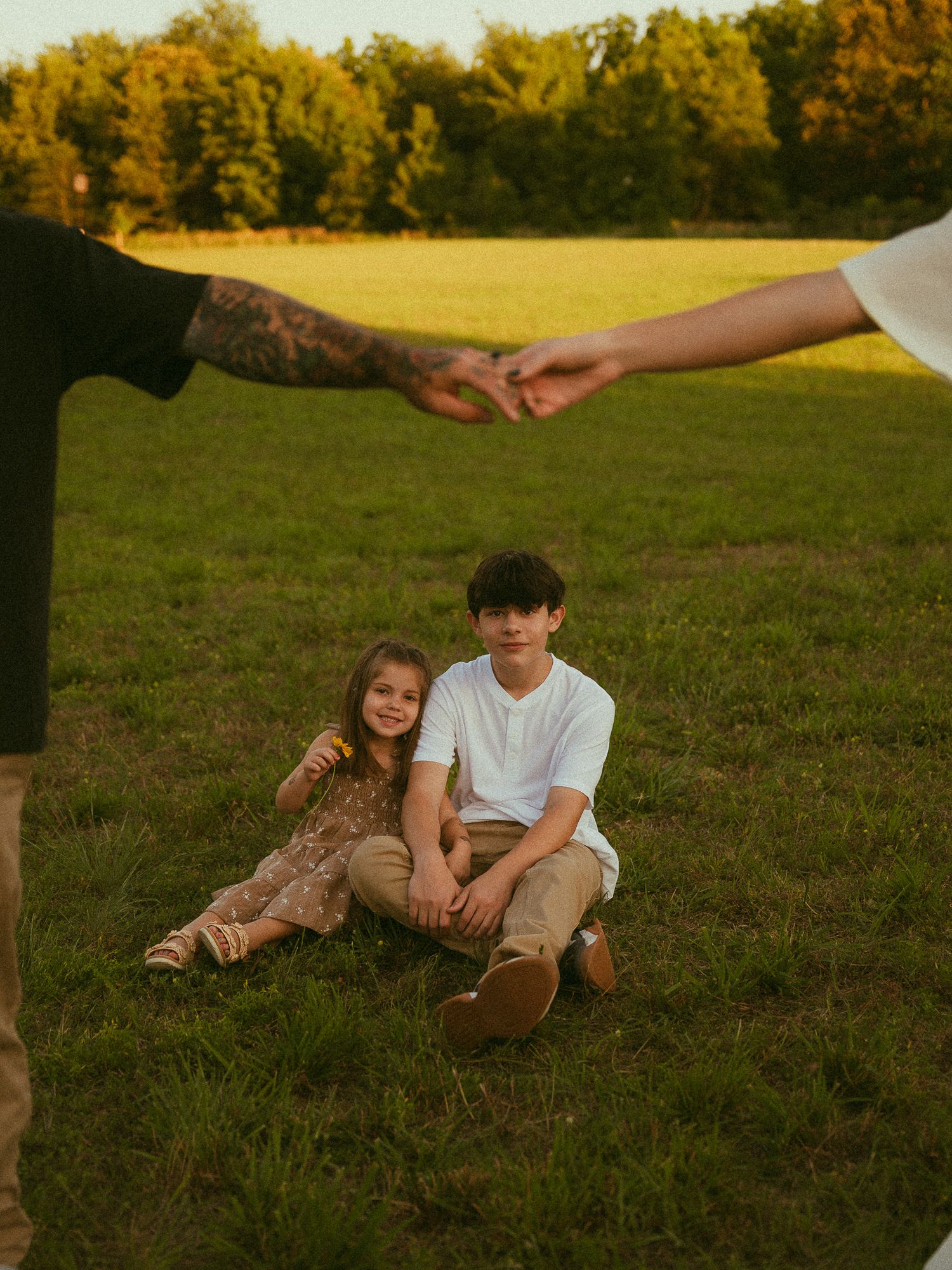 Two children sit on green grass while adults reach toward each other overhead during a golden hour family photo session.