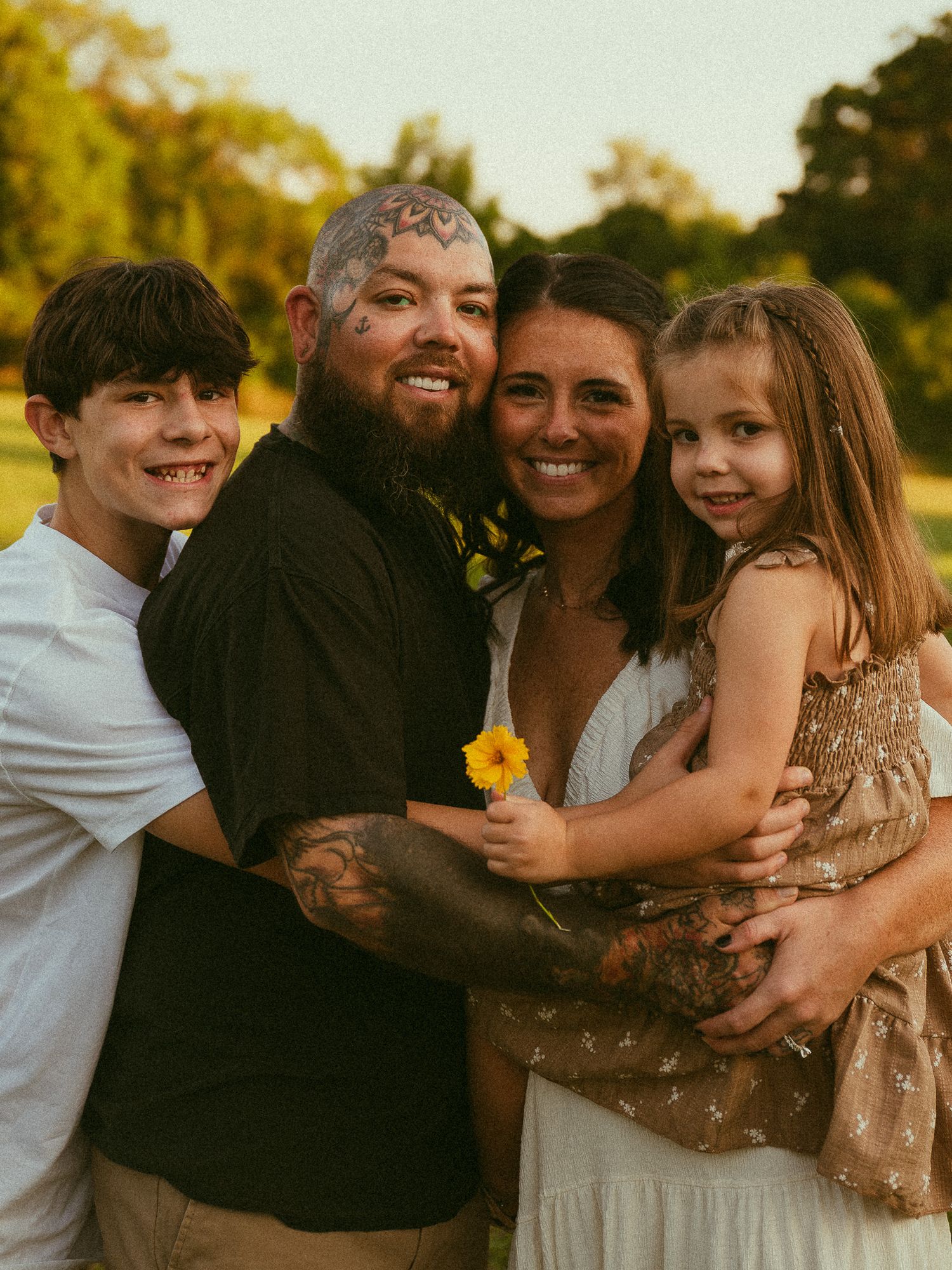 A tattooed man and woman smile with two children outdoors during golden hour in a warm family portrait.