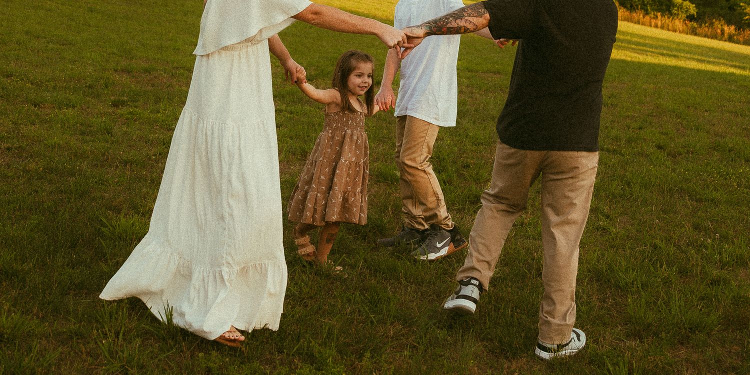 A family holds hands swinging a young girl in a floral dress on a green lawn at golden hour.