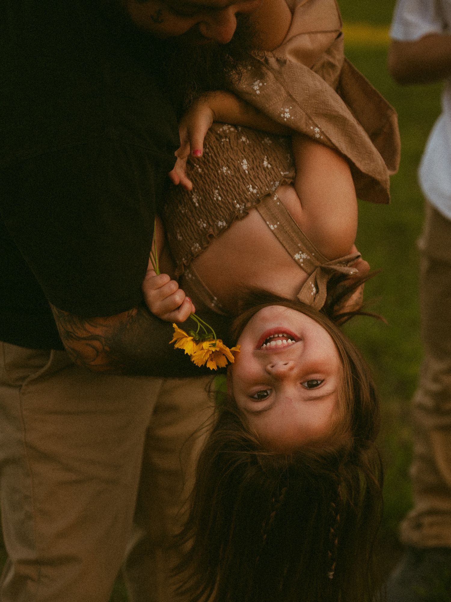 A laughing child held upside down by an adult, clutching a yellow sunflower in a sunny outdoor setting.