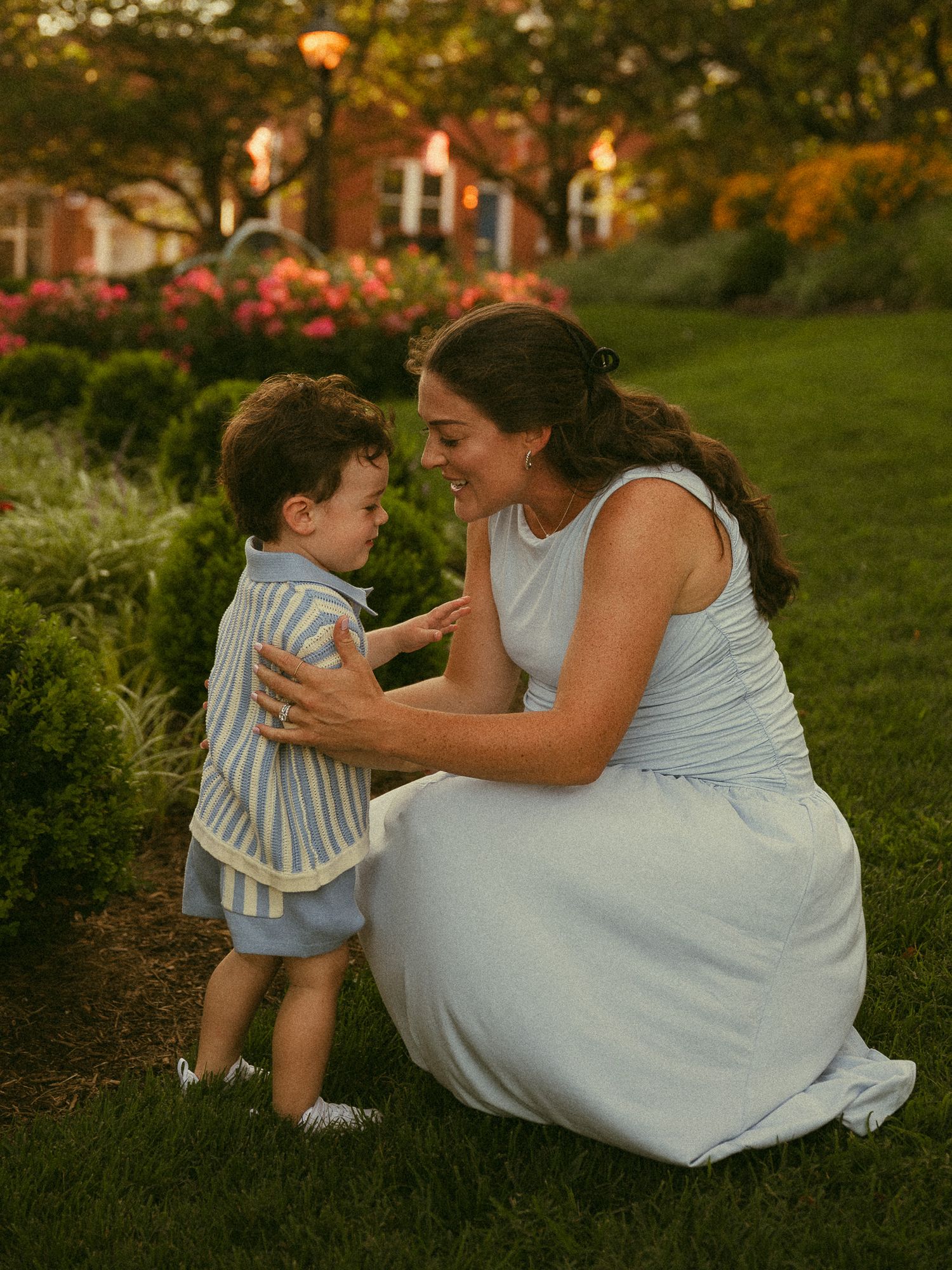 A mother in a white dress crouches on green grass, smiling at her toddler in a striped outfit at golden hour.