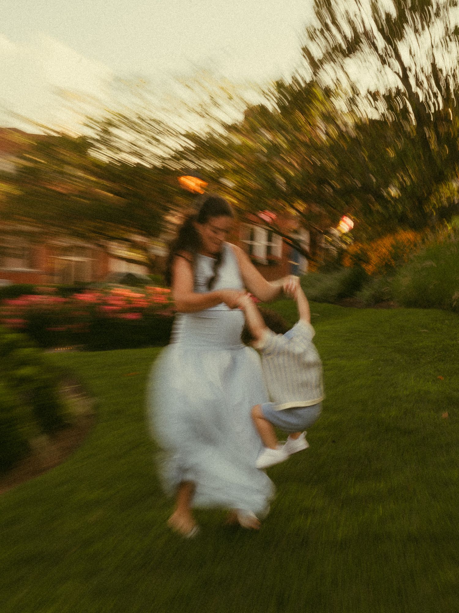 A woman in a blue dress joyfully swings a young child in a lush green garden at dusk.